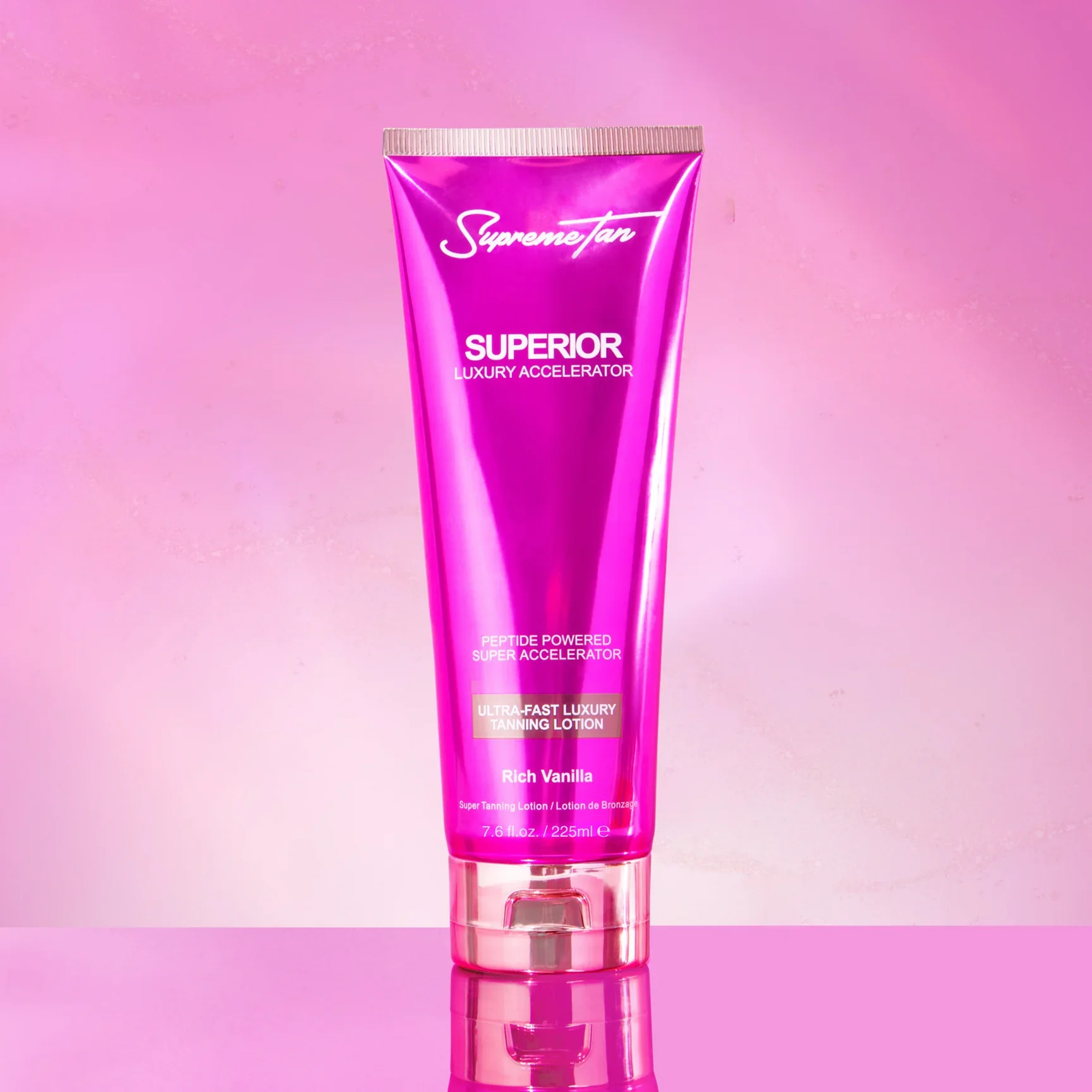A pink tube of SupremeTan Superior Luxury Accelerator tanning lotion with Rich Vanilla scent, standing on a reflective surface against a pink background.