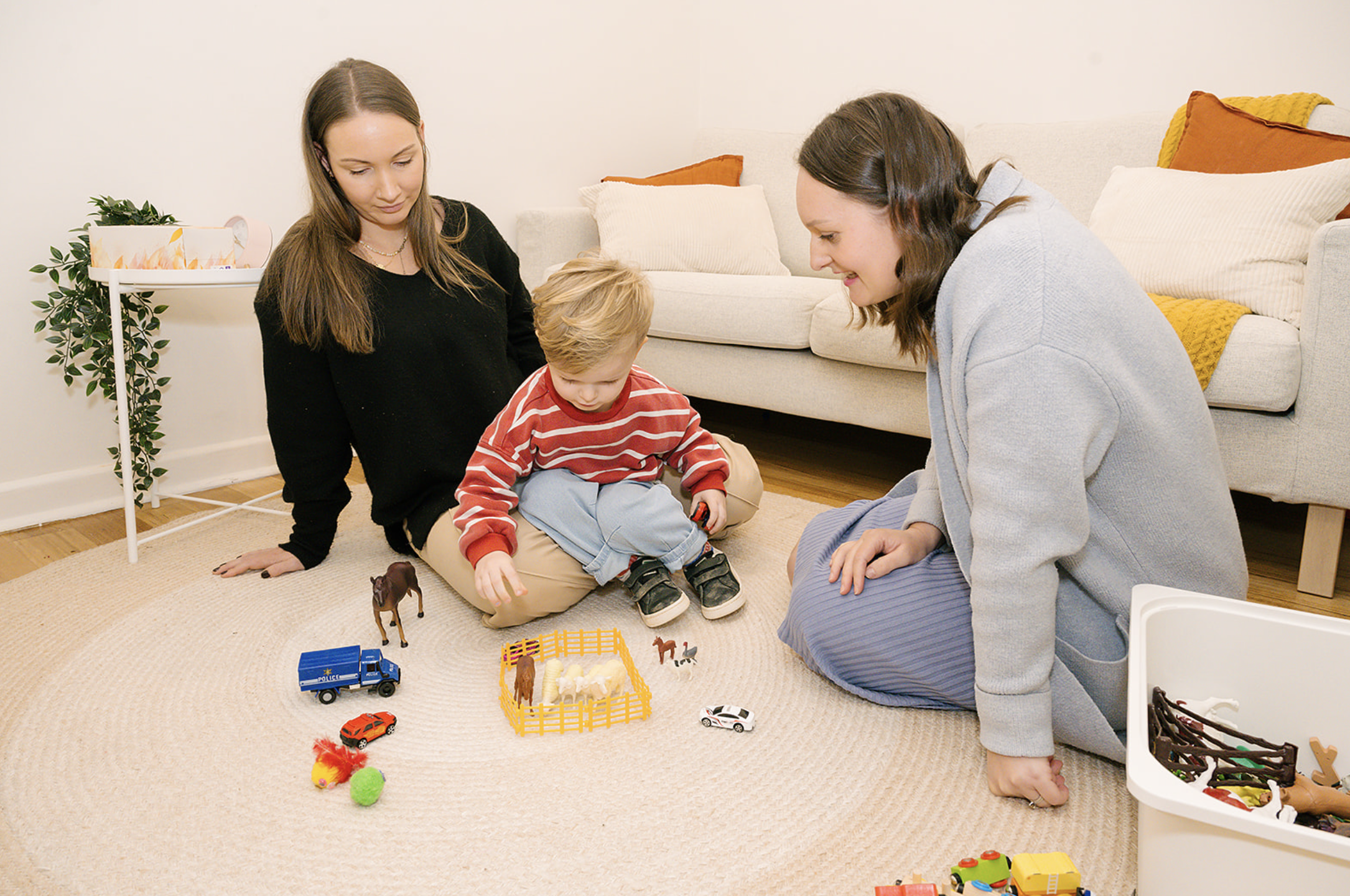 Two women and a young boy playing with toy cars and animals on a beige carpet in a living room with a white sofa and orange and yellow pillows.