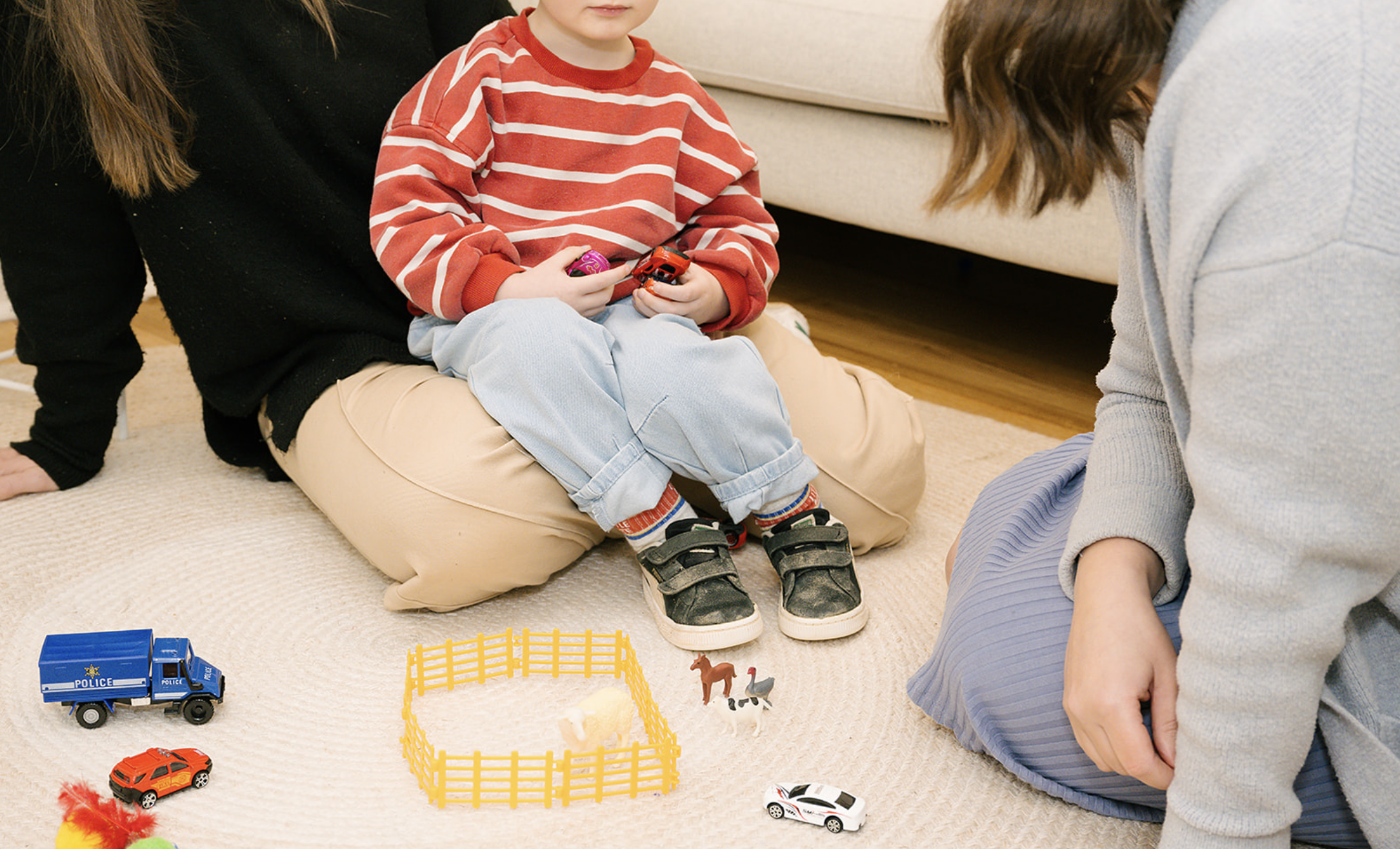 Children playing with toy cars on a beige carpet in a living room, with a woman sitting nearby and a small child sitting on her lap, holding a toy car.