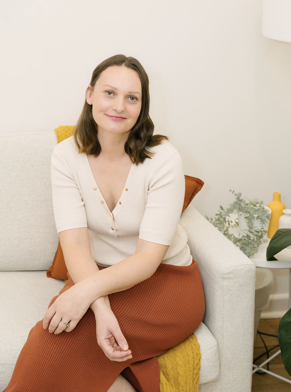 A woman with shoulder-length brown hair and a light complexion sitting on a beige sofa with yellow and brown cushions, smiling at the camera. She is wearing a cream-colored top and an orange skirt, with a gold ring on her finger. The background shows a white wall, a side table with decorative vases, and greenery.