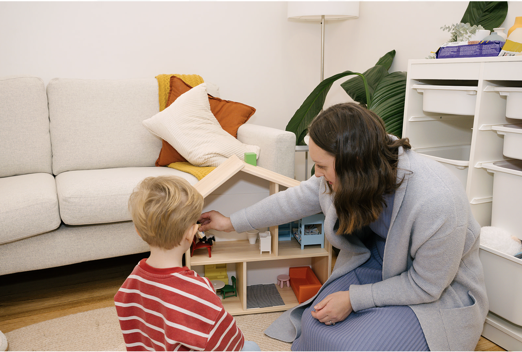 A woman and a young boy playing with dollhouse furniture in a living room. The woman is kneeling, and the boy is sitting on the floor, both engaged with the toys.