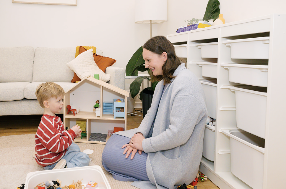 A woman and a young boy playing with toys indoors. The woman is sitting on the floor, facing the boy, and smiling. The boy, wearing a red and white striped shirt, is sitting on the floor with toys in front of him. There is a small dollhouse behind them, a couch with pillows in the background, and storage bins along a white shelving unit.