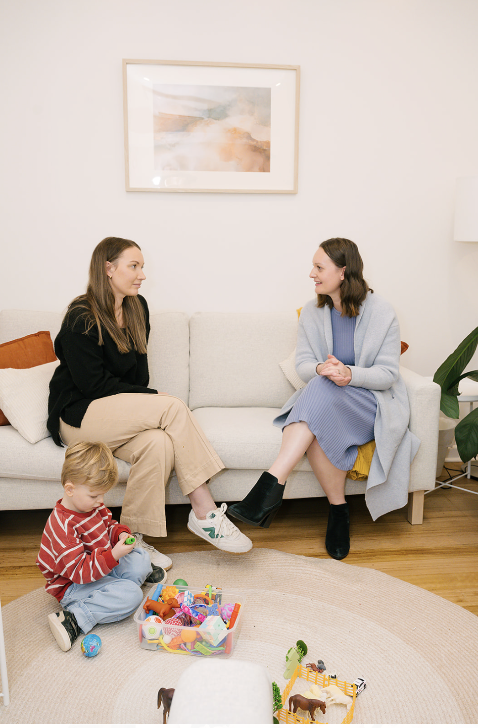 Two women sit on a beige sofa talking to each other, with a young boy playing on the floor nearby. The room has a minimalist decor with a framed painting, a potted plant, and toys on a rug.