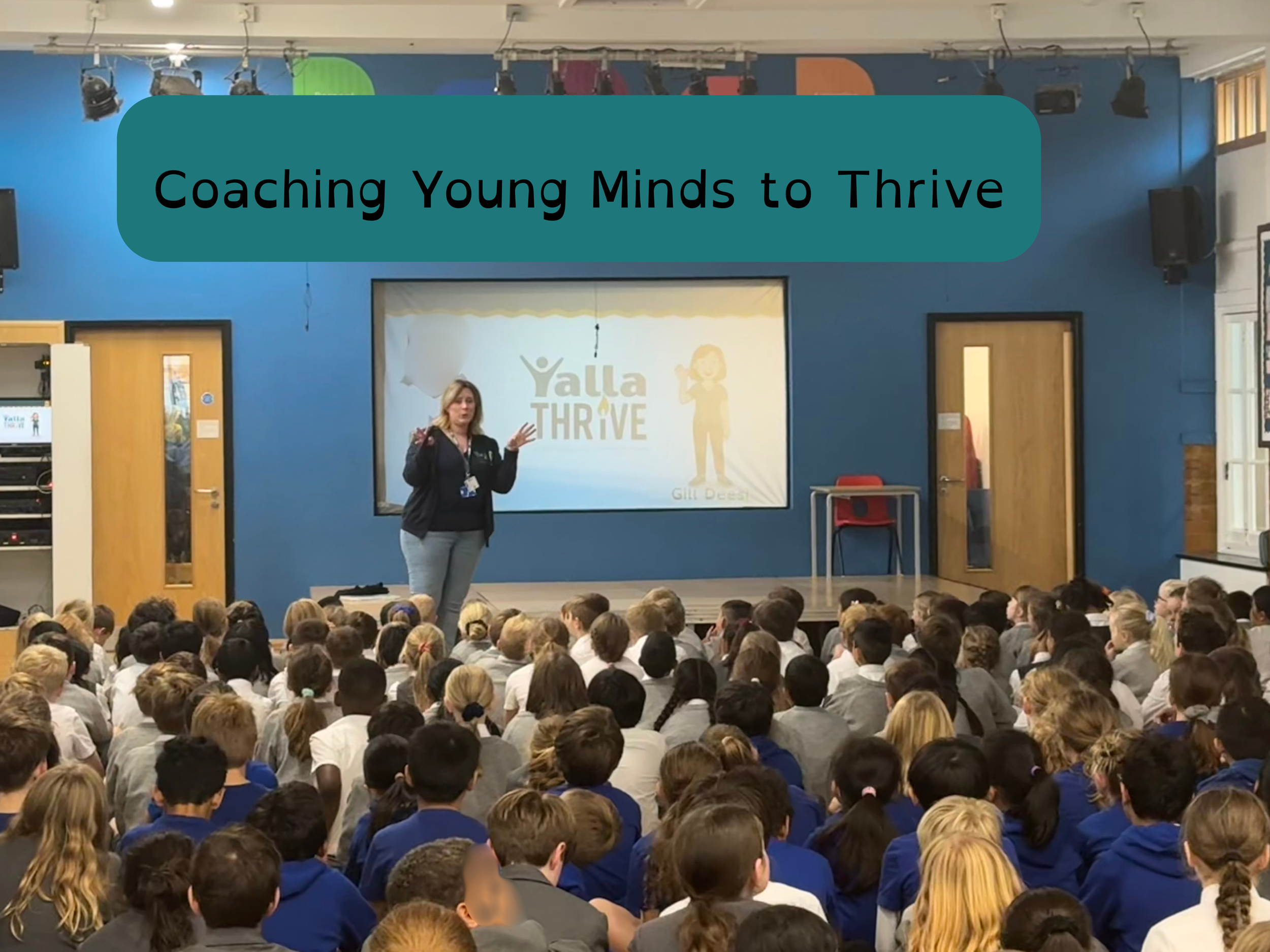 A woman giving a presentation to a large group of students seated on the floor in a school auditorium. The presentation screen displays the words 'Yalla Thrive' and a logo.