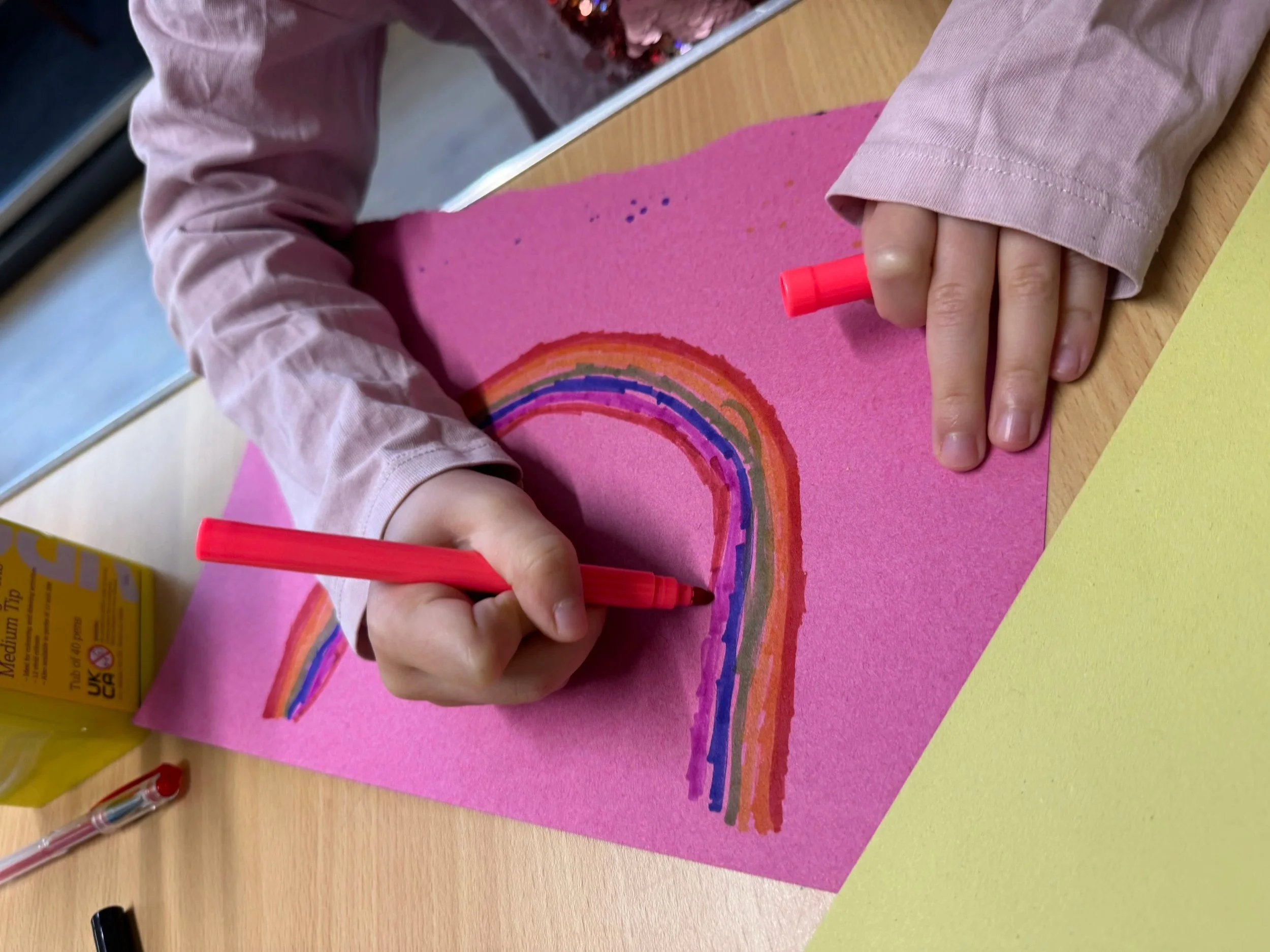 Child appears to be coloring a rainbow on pink paper with purple, orange, red, and other colors, using a pink crayon.