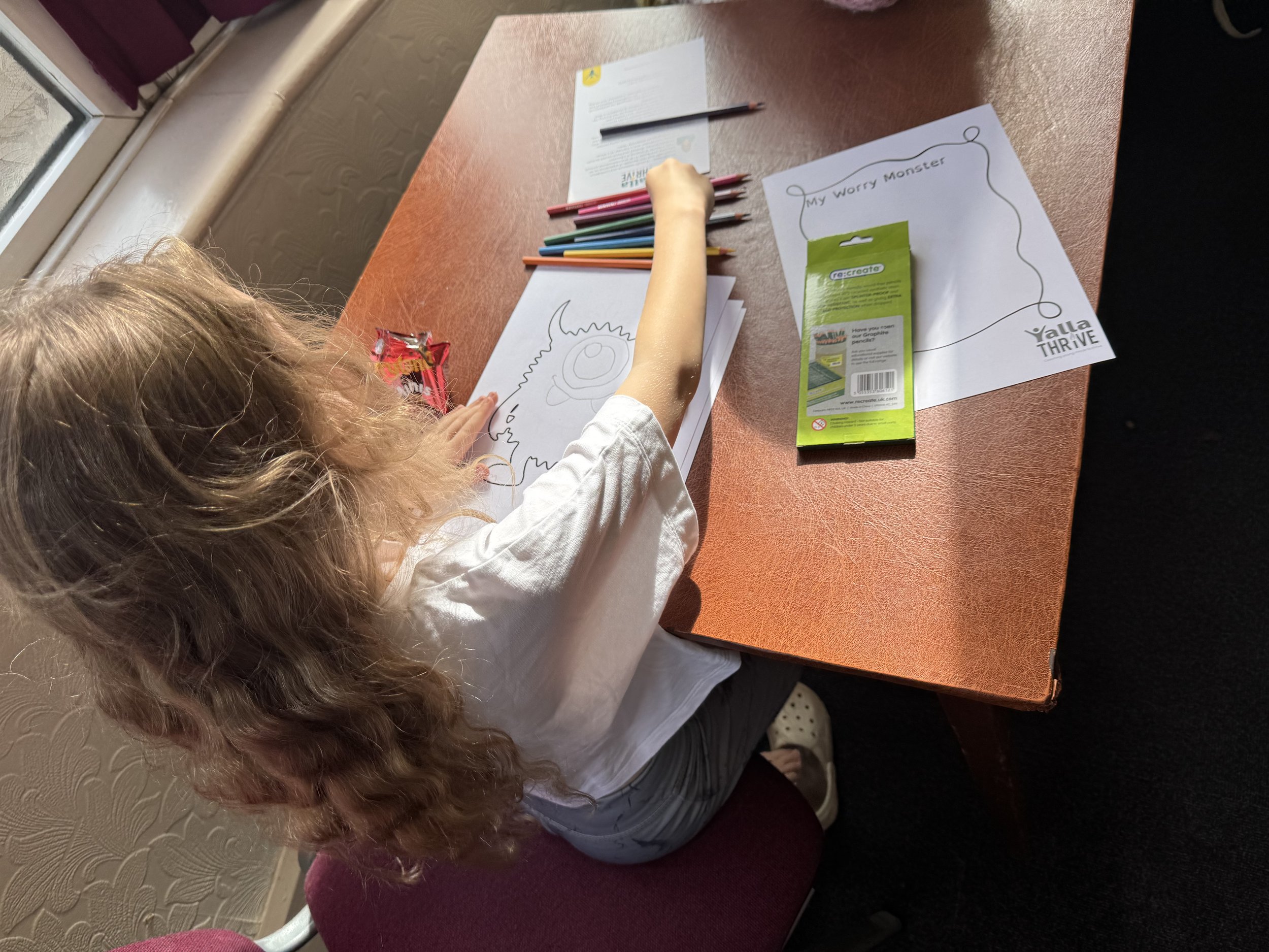 A child with curly hair drawing a monster on a piece of paper with colored pencils. There are also papers and a pack of graphite pencils on the table. The child is sitting on a red chair near a window.
