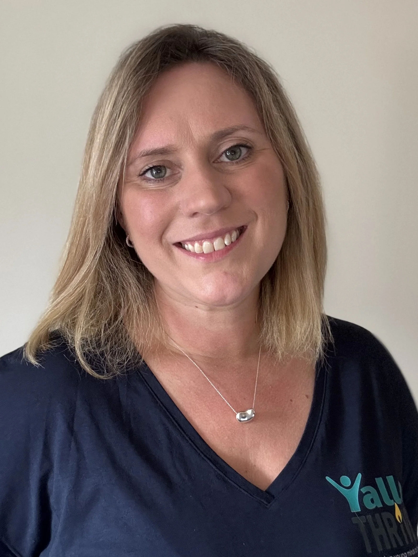 A woman with light brown hair and hazel eyes, smiling, wearing a dark-colored t-shirt with a logo and a silver necklace with a small charm, standing against a plain light-colored wall.
