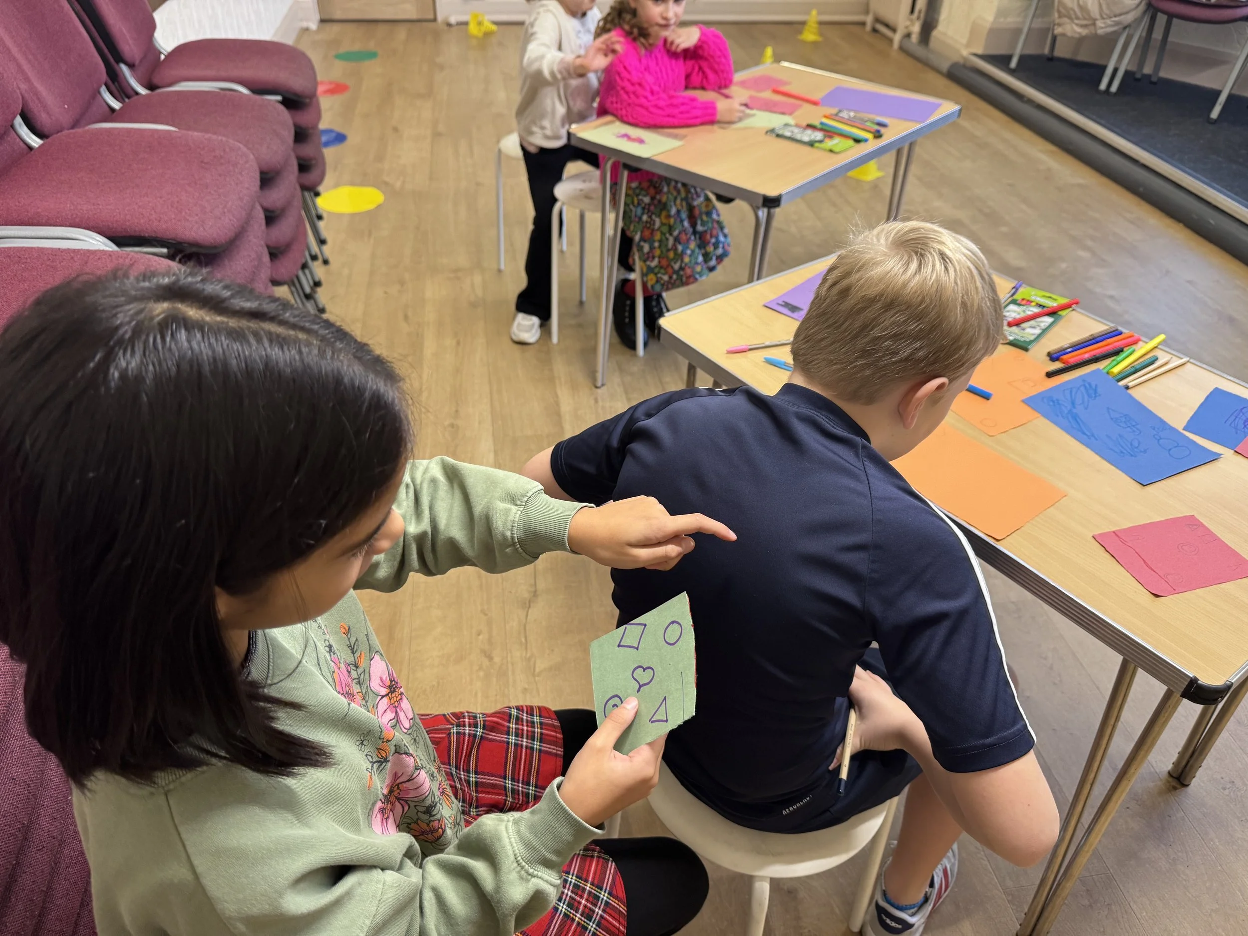 Children participating in a classroom activity, sitting at tables with colorful papers, markers, and educational materials, with some sitting and others reaching for supplies.