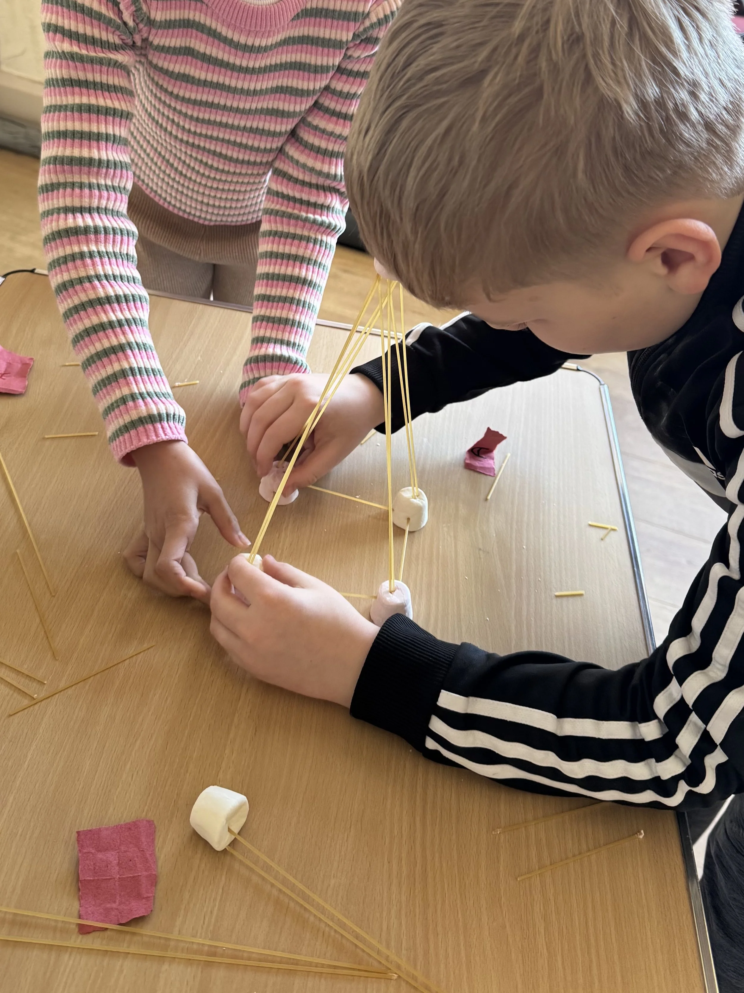 Two children working together on a project involving marshmallows, spaghetti sticks, and tissue paper on a wooden table.