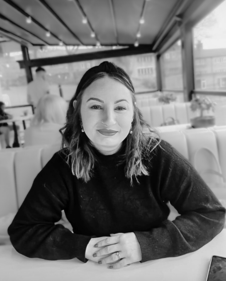 A woman with shoulder-length hair and earrings sitting at a table in a restaurant or cafe, smiling at the camera, with windows and other patrons in the background.