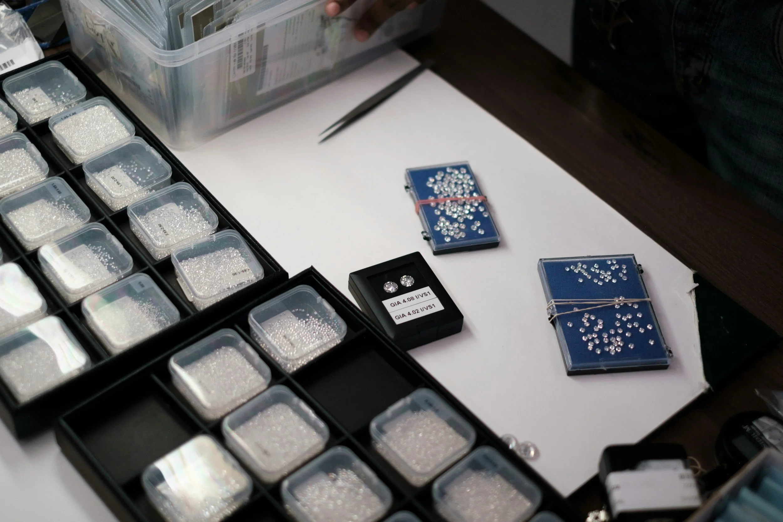 Assorted loose diamonds and gemstones in small plastic containers and on blue display trays, some in a small box, on a white surface.