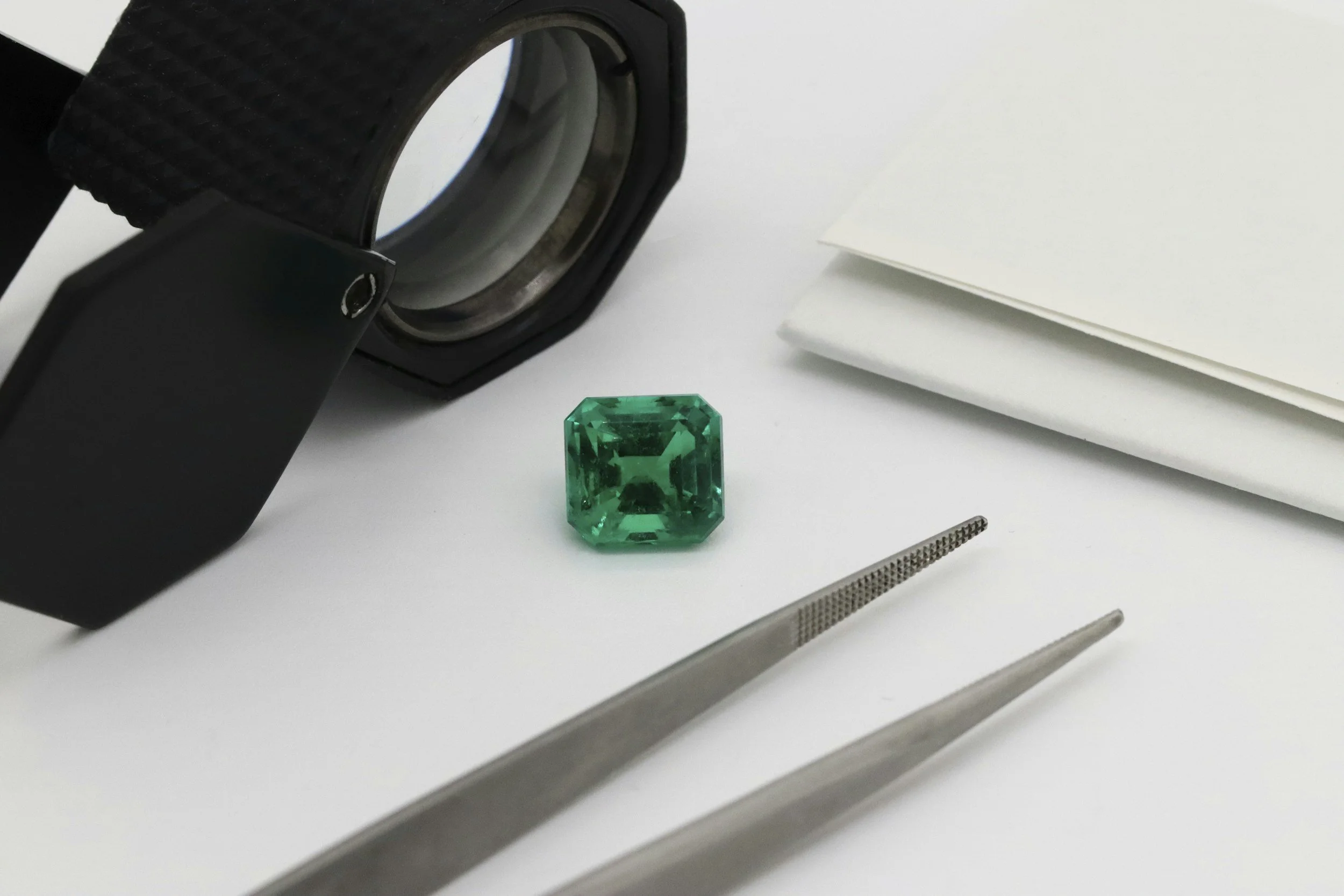 A small square-cut green gemstone resting on a clean white surface, surrounded by jeweler’s tools including tweezers and a loupe, with a notebook partially visible in the background.
