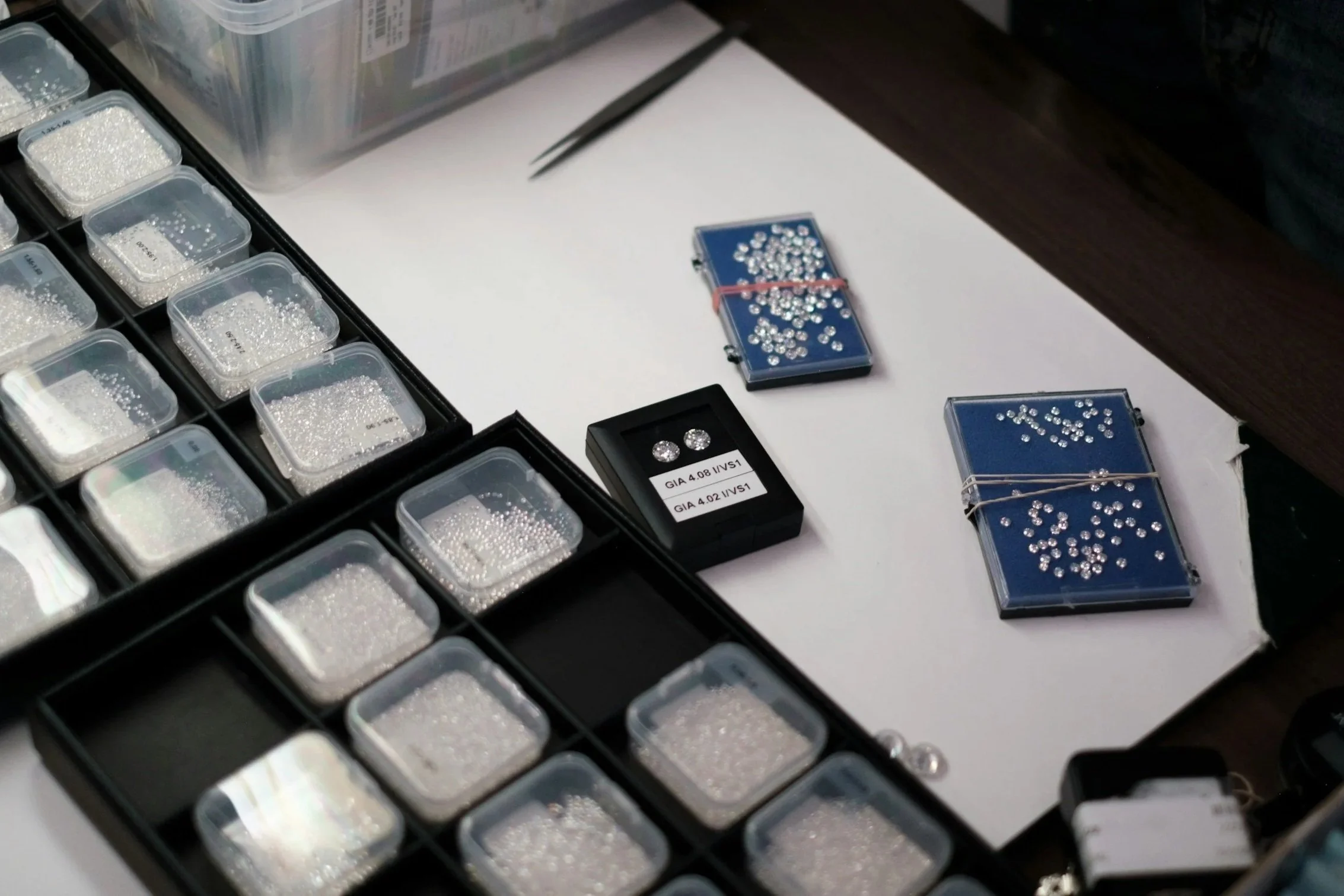Assorted loose diamonds and gemstones in small plastic containers and on blue display trays, some in a small box, on a white surface.