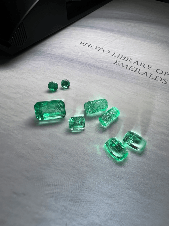 Assorted emeralds in various cuts, including faceted and carved stones, displayed on a light surface with soft green reflections.