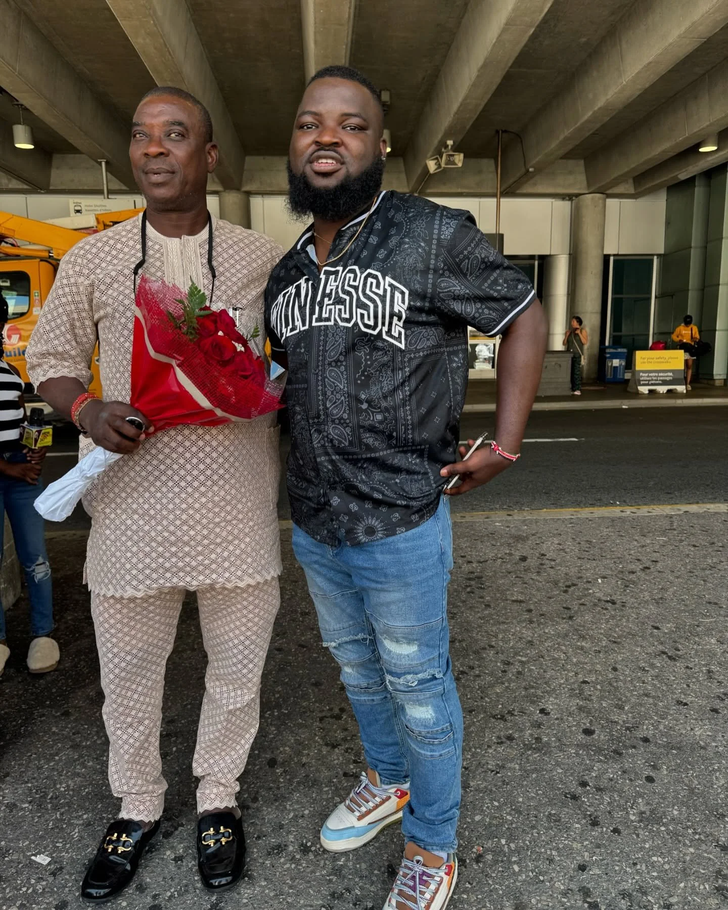 Two men standing under an overpass, one holding a bouquet of red roses, dressed in traditional attire, and the other wearing a black shirt with white lettering and distressed jeans, posing for a photo.