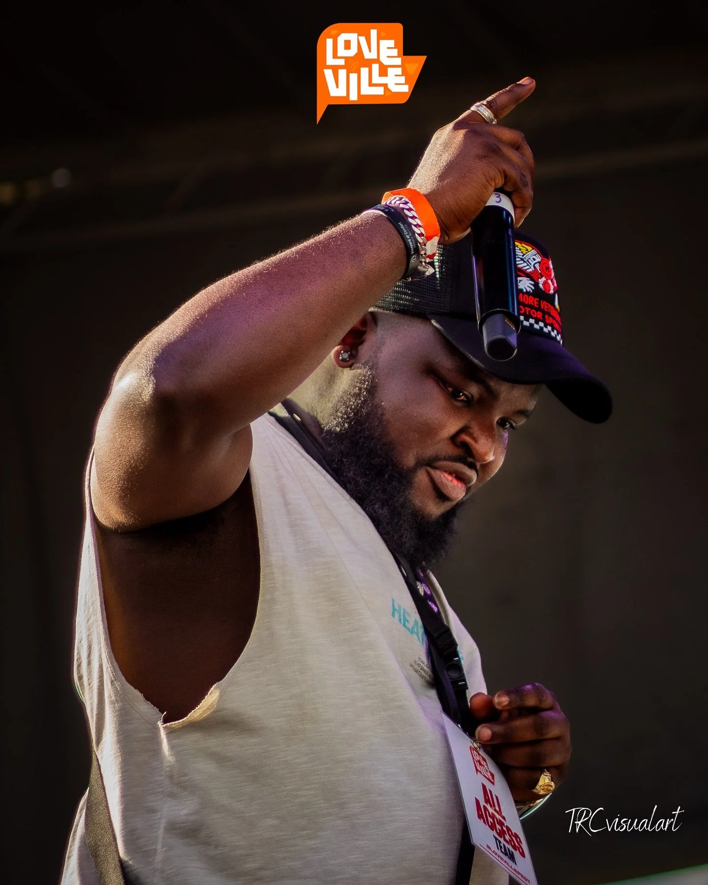 Close-up of a man with dark skin holding a microphone in his right hand, wearing a black cap, a white sleeveless shirt, silver chains, and a red wristband. The background is black, and there is an orange and white logo reading 'Love Ville' in the top