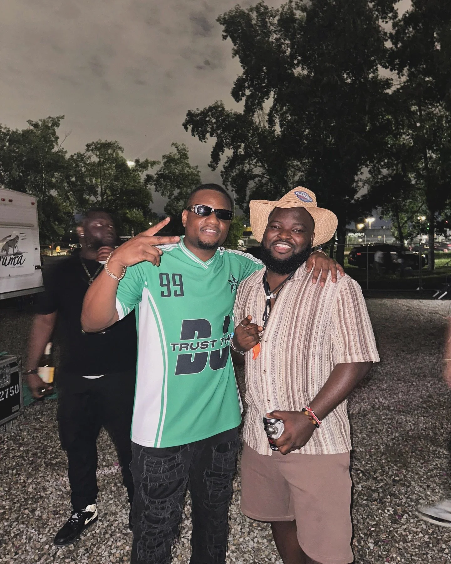 Two men smiling and posing with their arms around each other outdoors at night. One wears a green sports jersey and sunglasses, making a peace sign. The other has a beard and wears a wide-brimmed hat and striped shirt, holding a bottled beverage.