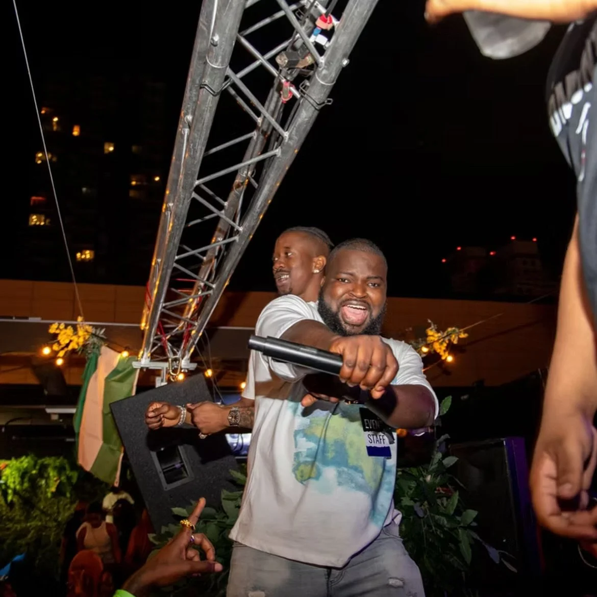 A man with a beard holding a microphone and pointing forward, smiling at the camera, with another man in the background. They are at an outdoor event at night, with stage equipment and decorative plants around.