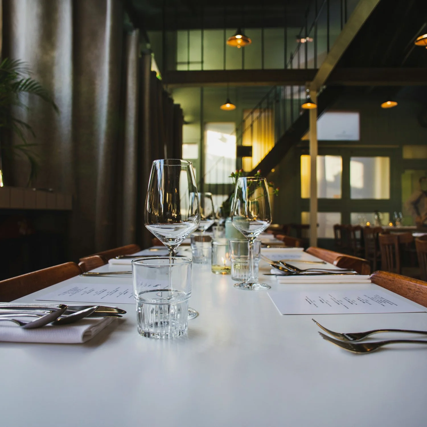 A restaurant table set with wine glasses, water glasses, plates, and silverware in a modern, well-lit dining room with wood accents and hanging lights.