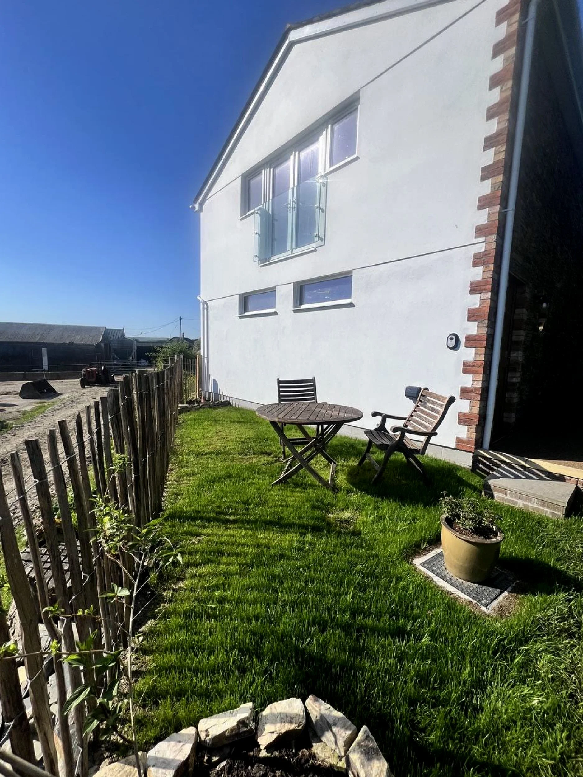 Backyard patio with a small round table and chairs, a potted plant, a grassy area, a wooden fence, and a white house with brick accents under a clear blue sky.