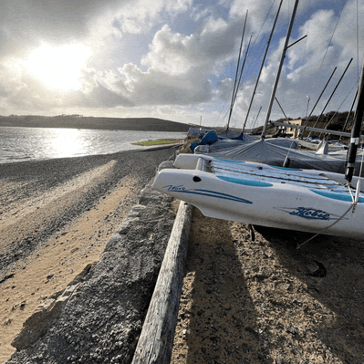 Sailboats on the shore next to a sandy beach and a body of water with hills in the background under a cloudy sky.