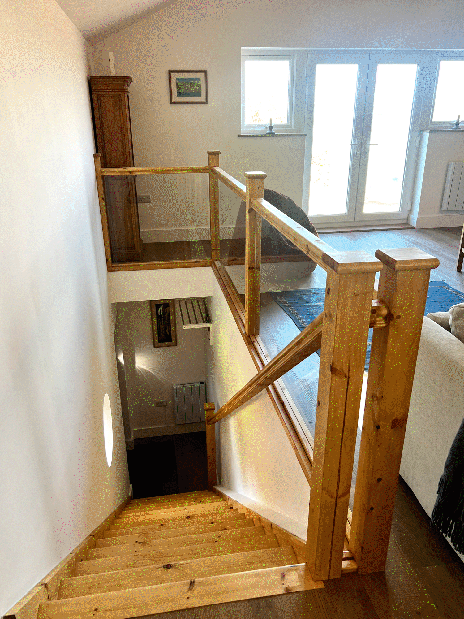 Inside view of a wooden staircase with a glass railing, beige walls, a small landscape painting, and large windows letting in natural light.