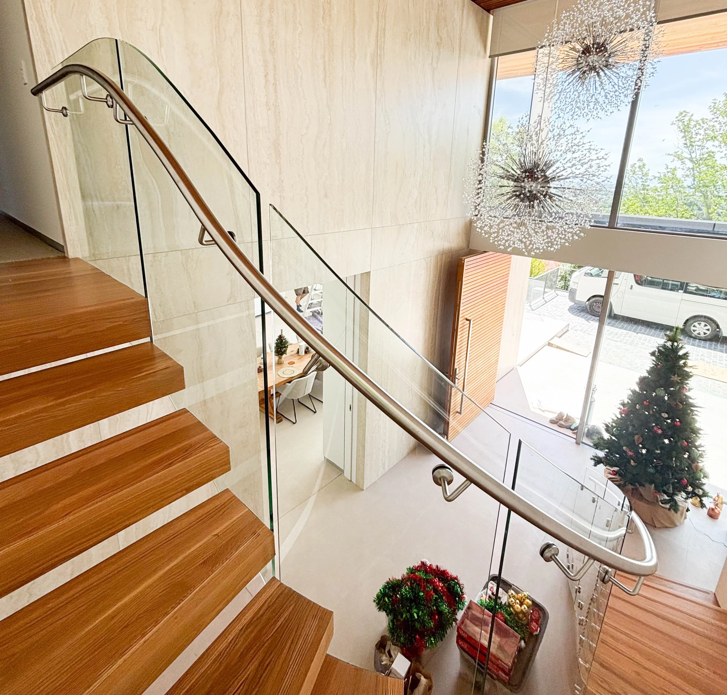 Indoor staircase with wooden steps, metal handrail, glass railing, Christmas decorations including a Christmas tree with ornaments and a gift basket, large window with natural light, and modern hanging light fixtures.