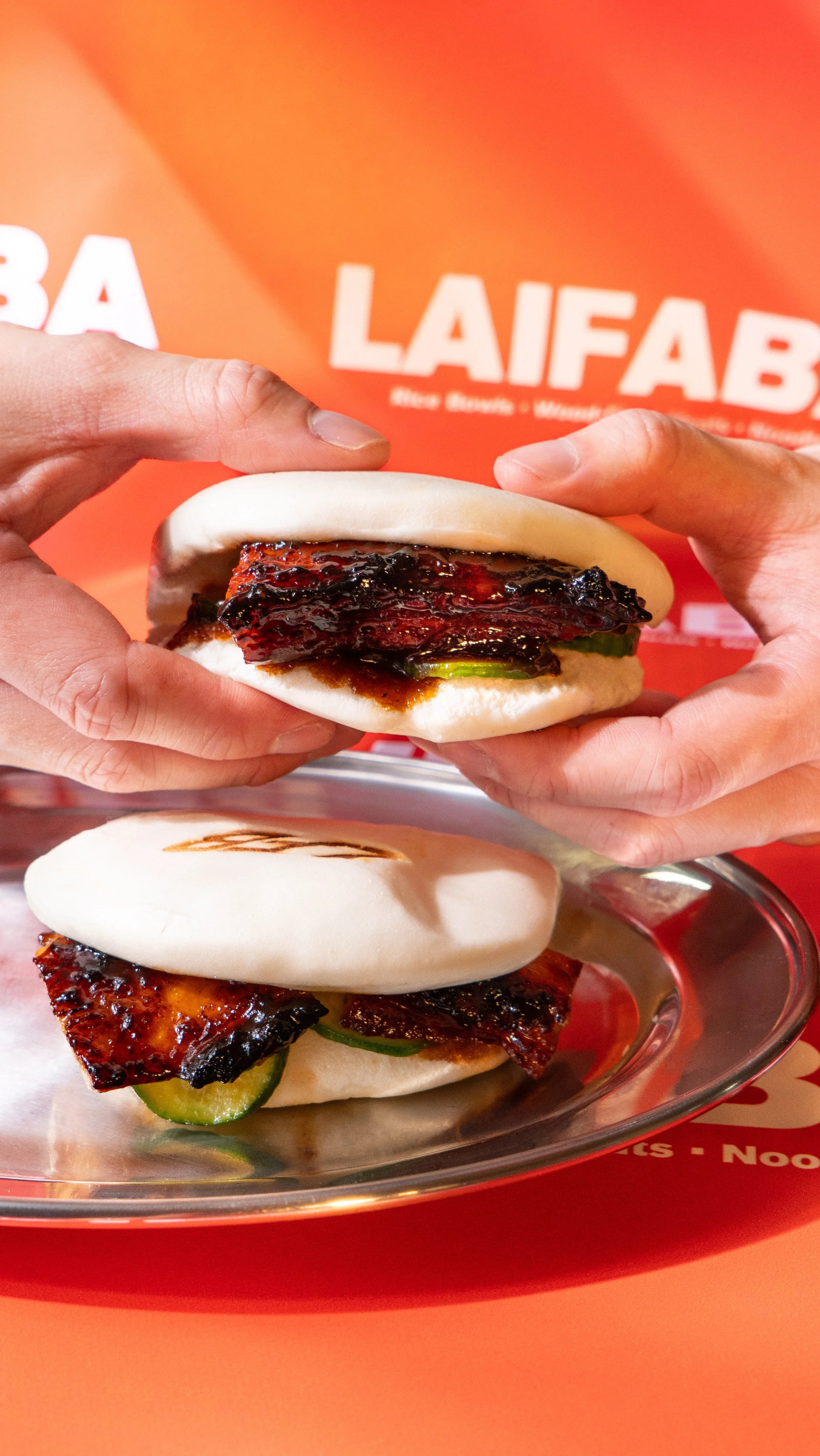Close-up of hands holding a steamed bun filled with barbecue pork, with another bun and barbecue pork on a metal tray in the background against an orange background with the word 'LAIFAB' visible.
