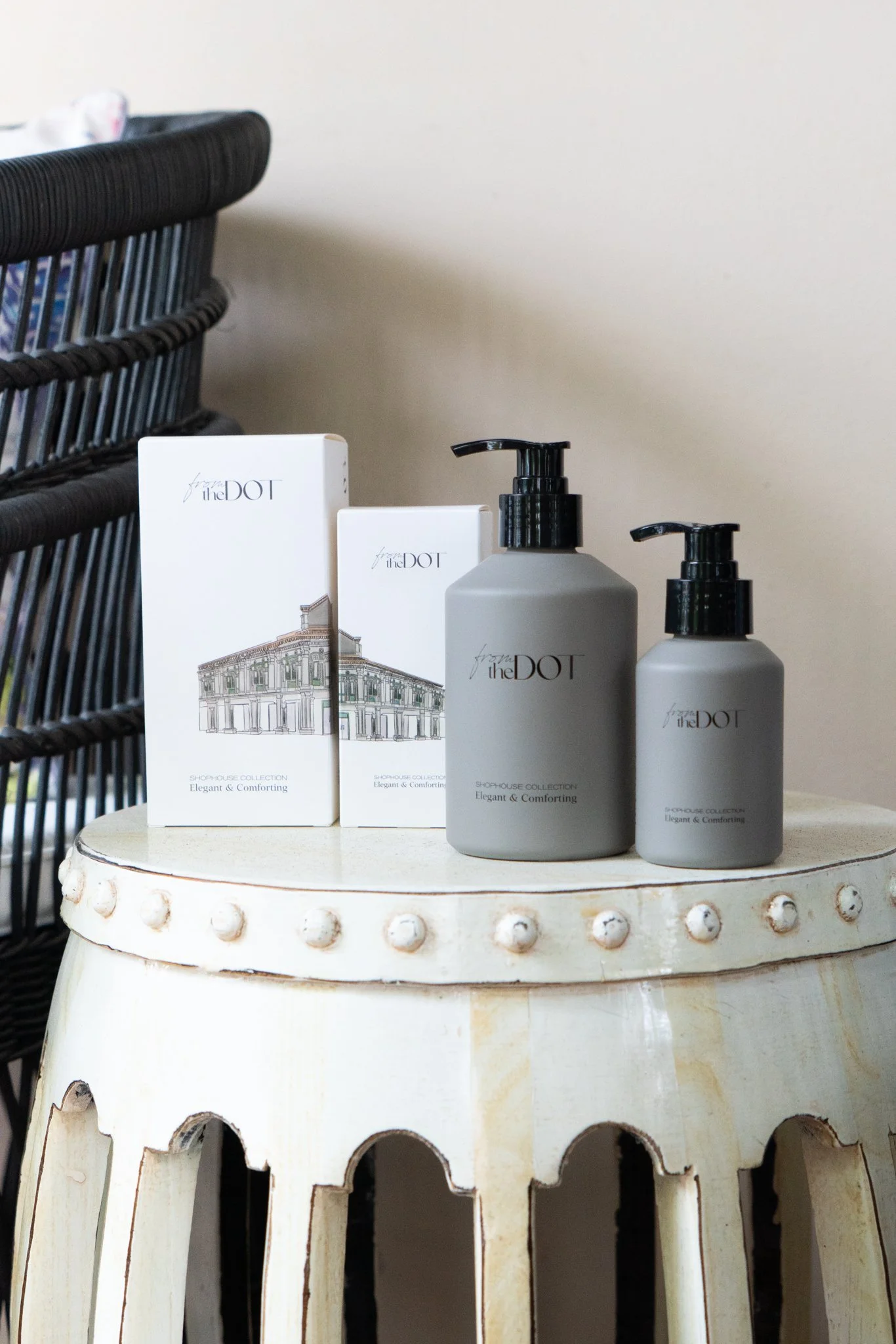 Grey pump bottles and boxes of The Dot soap or lotion on a worn white side table with decorative edge, beside a black wicker chair and a plain wall.
