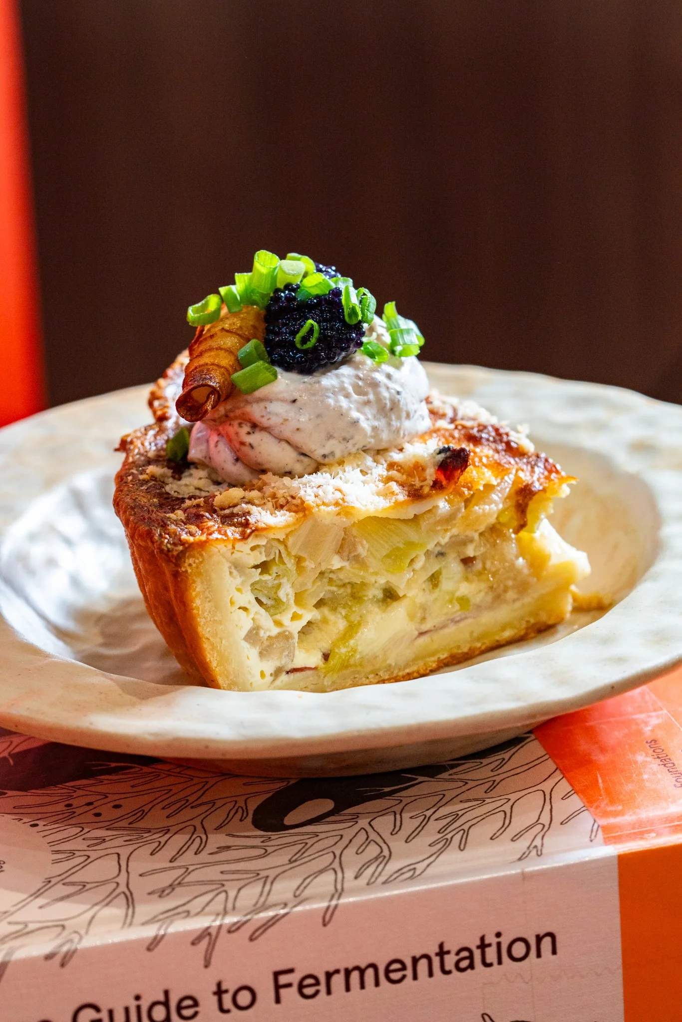 A savory quiche with a crispy crust filled with cheese, onions, and other ingredients, topped with sour cream, chopped green onions, a black caviar dollop, and a rolled piece of ham in a white ceramic dish.