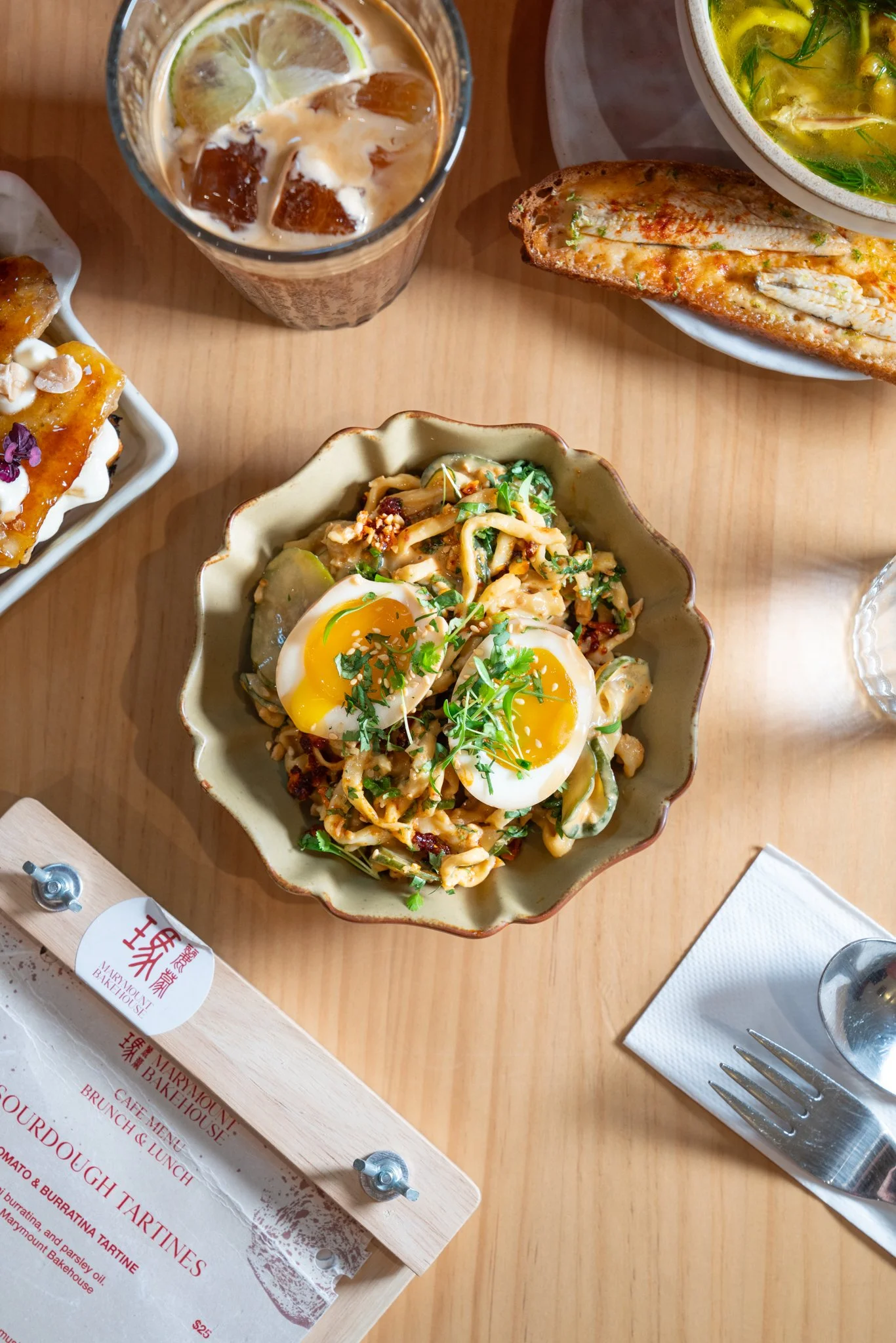 A bowl of noodles topped with soft-boiled eggs and garnished with herbs, a glass of iced beverage with lemon, a bowl of soup, a plate of grilled fish, and a chocolate dessert on a table.