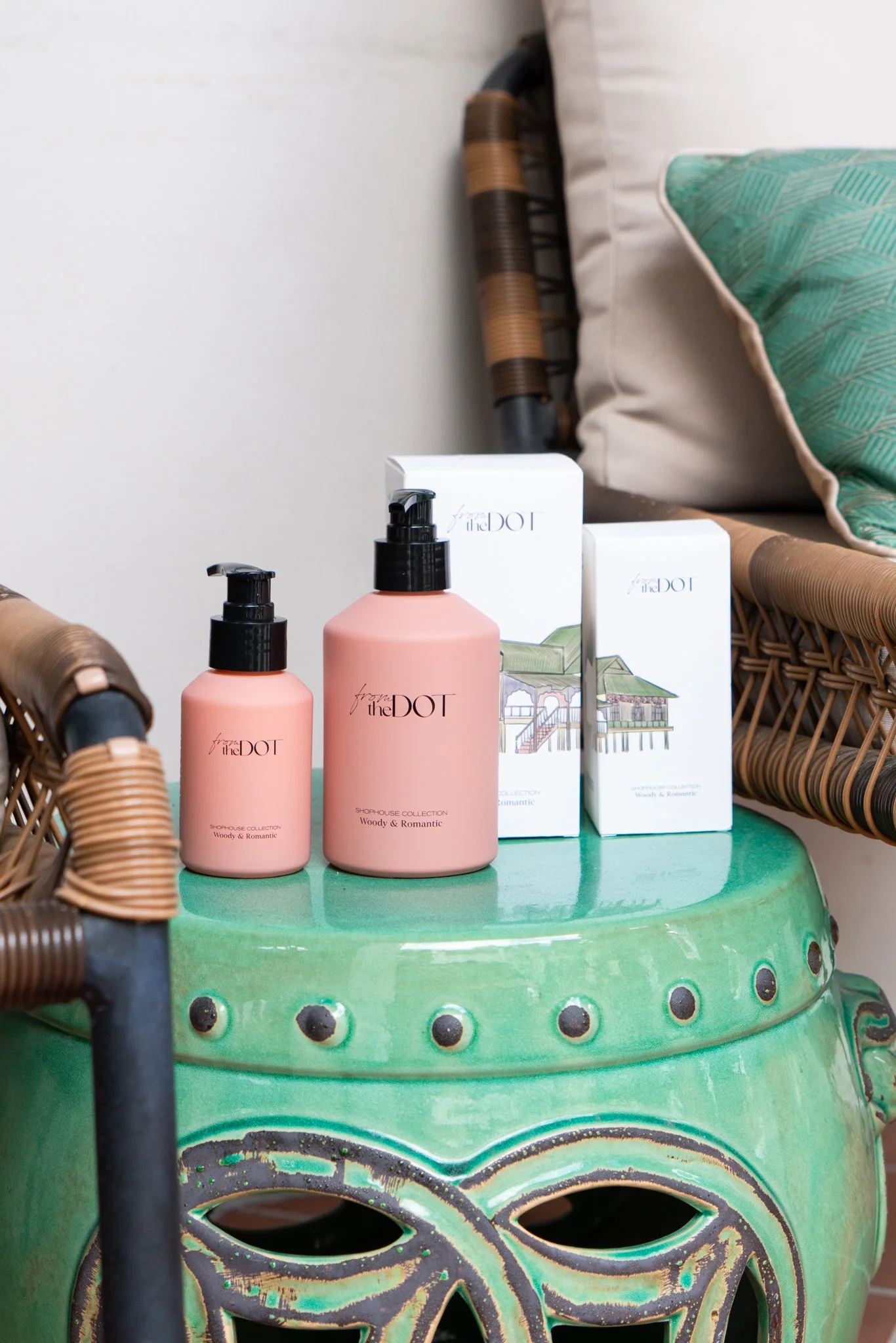 Pink bottles and boxes of From the Dot skincare products on a green ceramic table next to wicker chairs and green pillows.