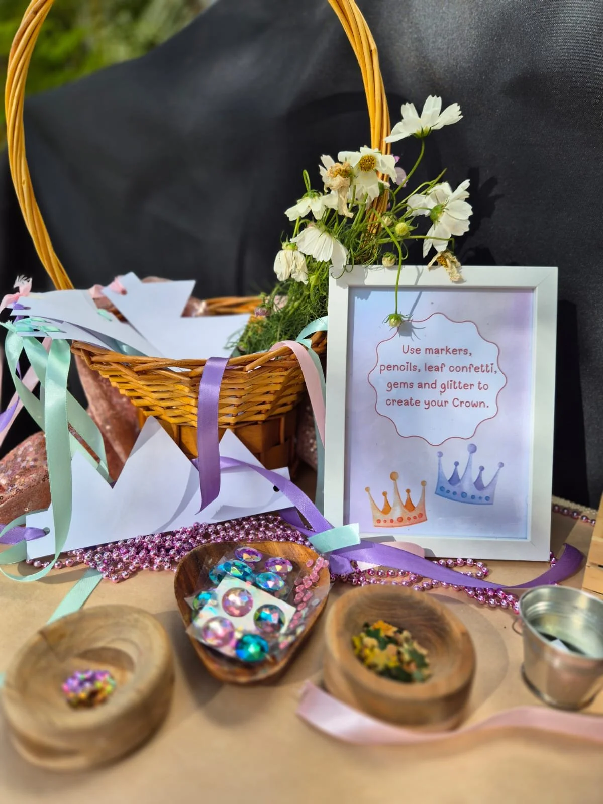 A decorated table with a wicker basket filled with paper, ribbons, and craft supplies, a framed sign with instructions to decorate crowns with markers, pencils, leaf confetti, gems, and glitter, and small wooden bowls containing decorated craft items