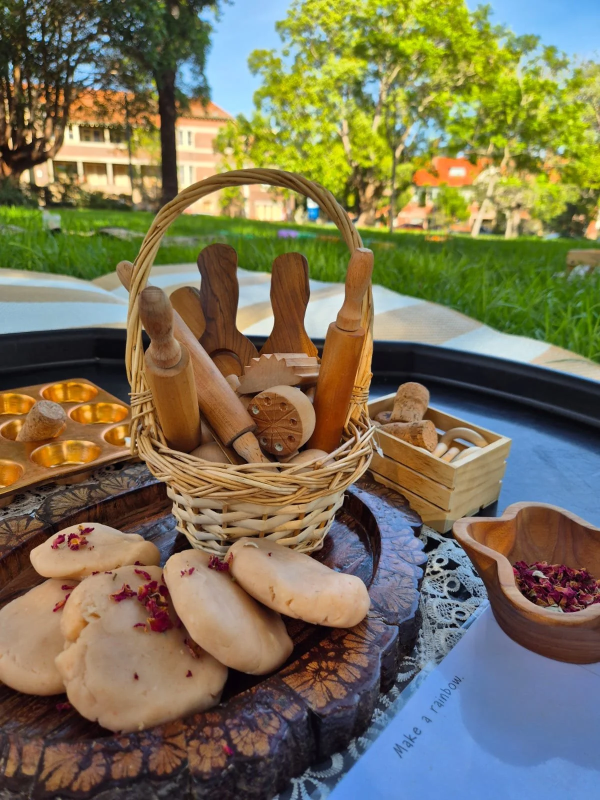 Cookies with pink petals on a wooden tray, a wicker basket filled with small wooden rolling pins and cookie cutters, a wooden box with cookie stamps, and a bowl of dried rose petals on a decorated table outside with green trees and buildings in the b