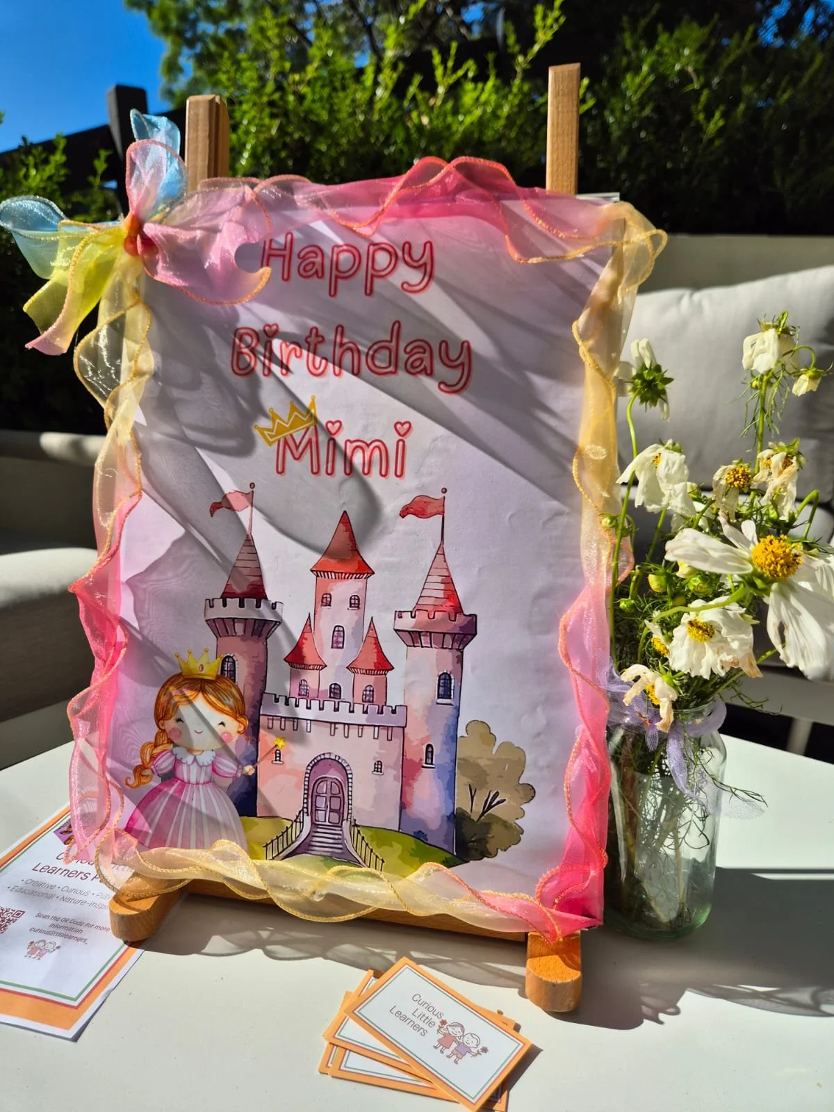 Colorful birthday card with a castle illustration and a fairy princess, decorated with pink and yellow ribbon, placed on a table near a glass jar of white flowers, under bright sunlight.