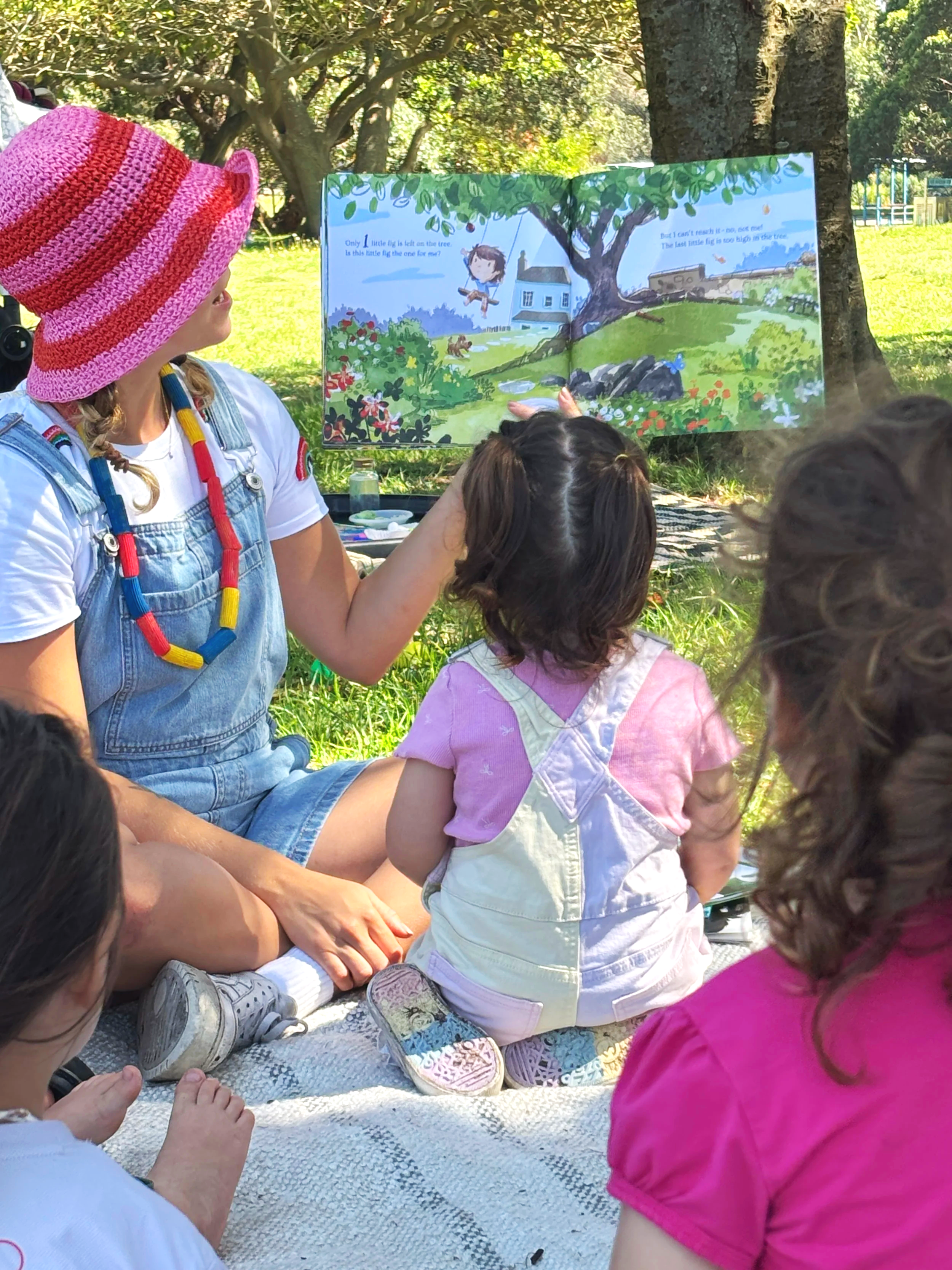 A group of children sitting on a blanket outdoors while an adult reads a colorful picture book to them. The setting is a sunny park with trees in the background.
