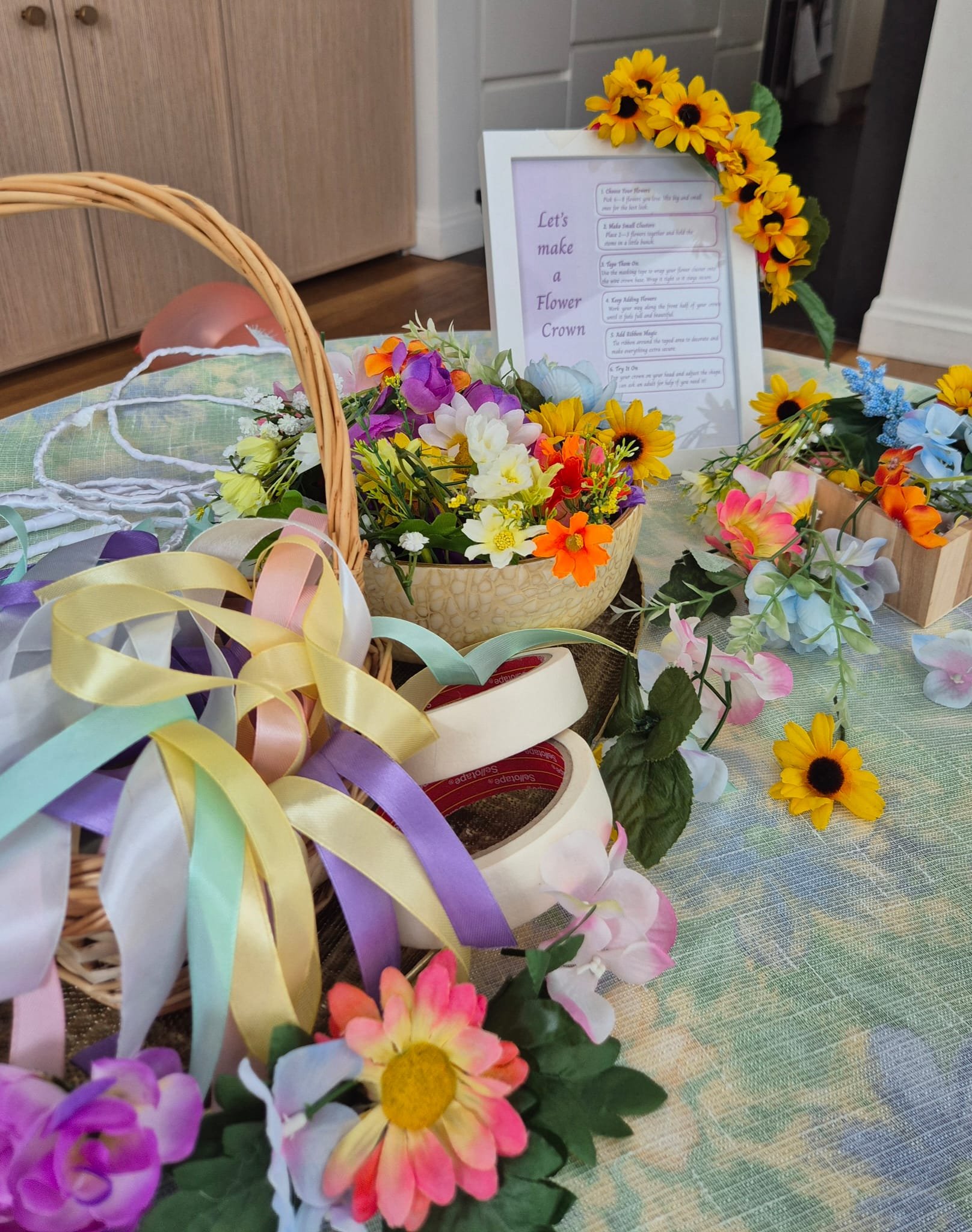 Colorful artificial flowers arranged for a flower crown craft, with a framed instructions guide and ribbons on a table.