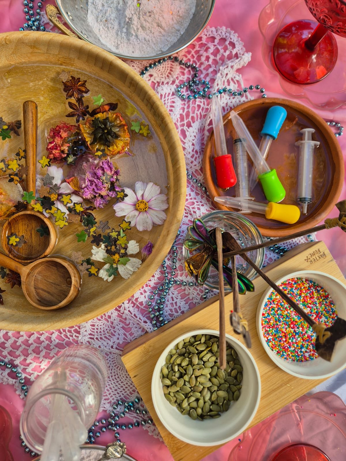 Colorful table setting with decorating supplies including flower petals, sprinkles, syringes, and beads, suggesting a craft party or celebration.