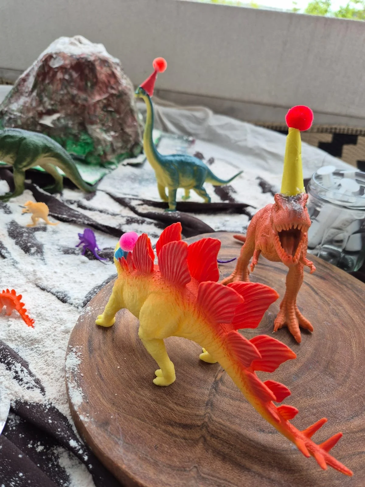Collection of toy dinosaurs on a wooden surface, with some wearing party hats, and a mountain model in the background.