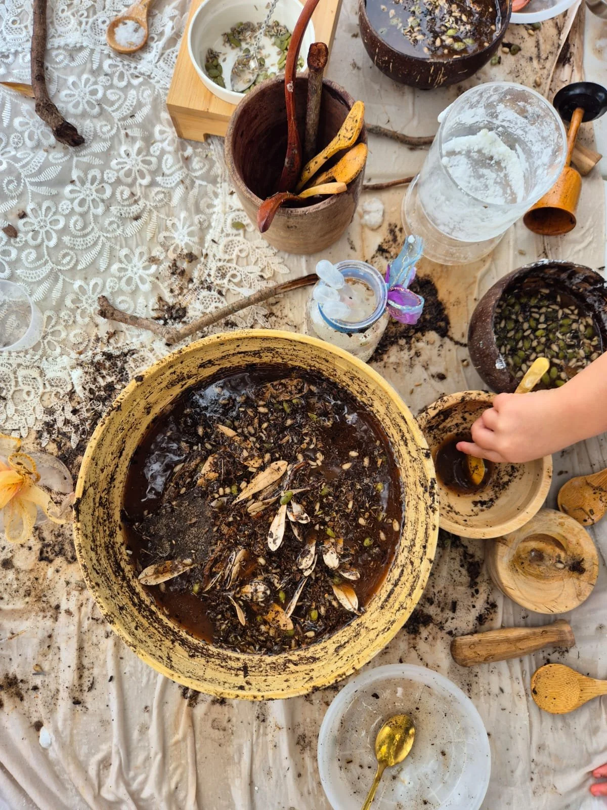 A messy table with dirt and plant debris, several containers holding soil, seeds, and possibly shells, a child's hand holding a small container, and various gardening tools, indicating a gardening or planting activity.
