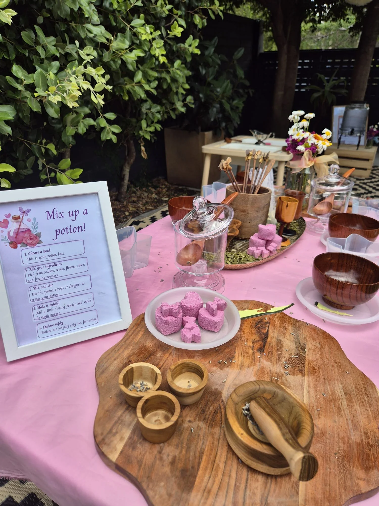 Table set for a potion-mixing activity with pink tablecloth, wooden bowls and spoons, pink marshmallows, flower arrangements, and a sign with instructions. Outdoors with trees and a black fence in the background.