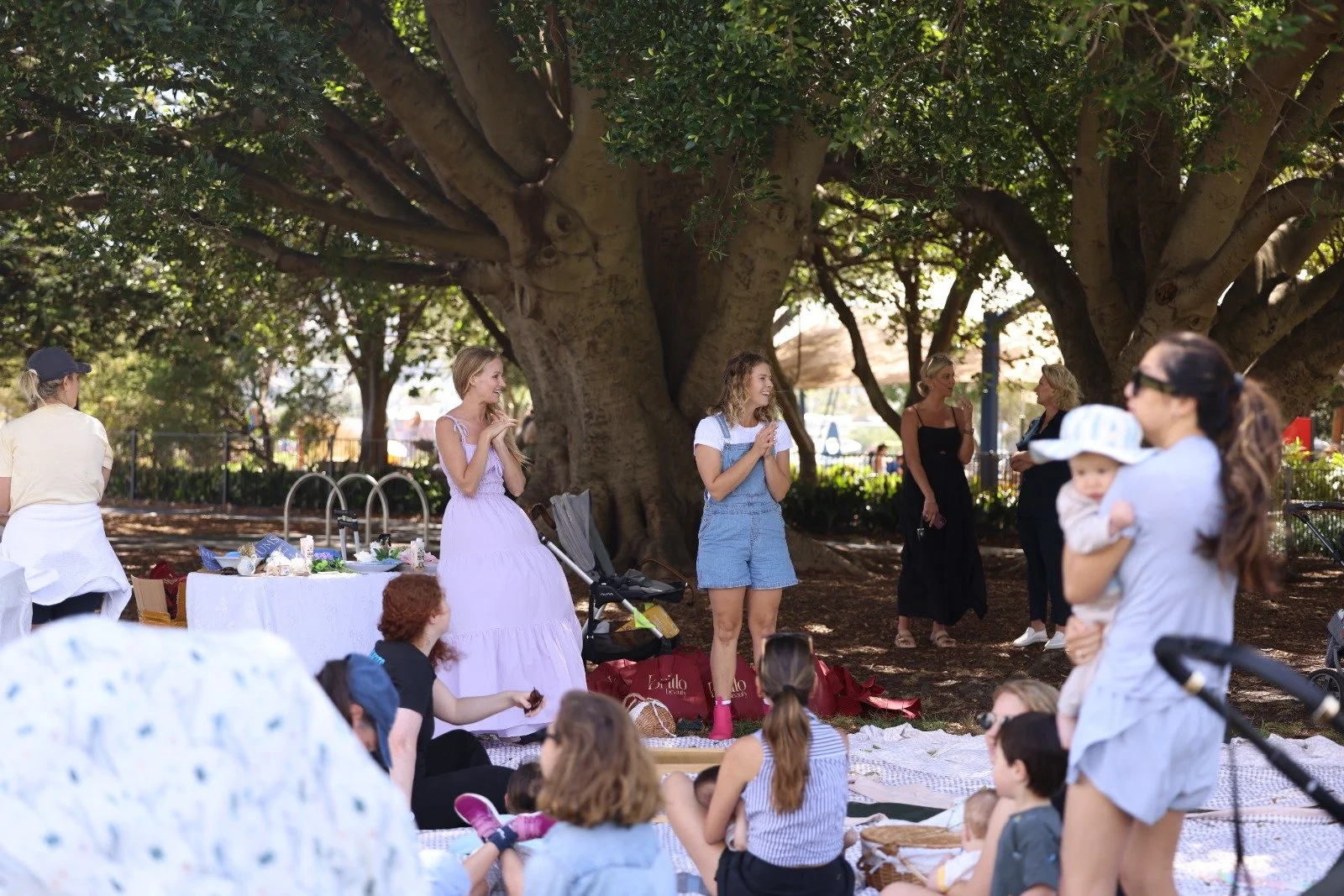 A group of women and children gathered outdoors under large trees, some sitting on blankets, with a few standing and talking. The scene appears to be a casual social event or picnic in a park during daytime.