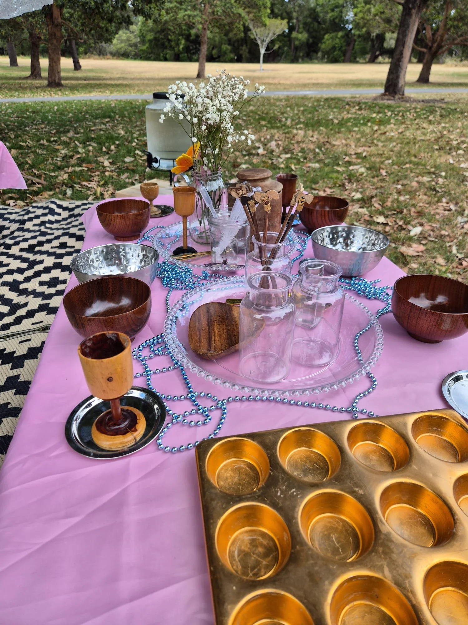 Table set with small wooden bowls, glass jars with flowers, blue beads, and a brass-colored mold with six round cavities, outdoors on a pink tablecloth surrounded by trees and grassy area.