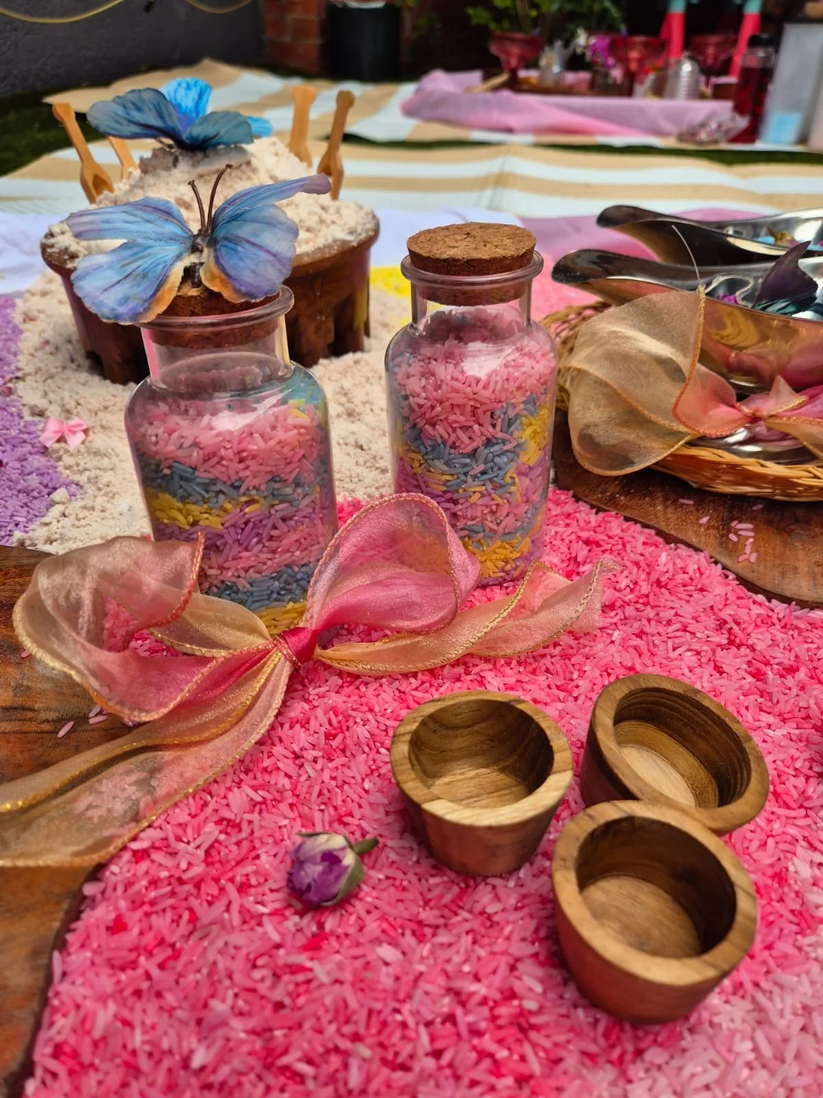Decorative setup with pink and multicolored rice, glass bottles with cork stoppers, wooden bowls, and butterfly decorations on a pink tablecloth.