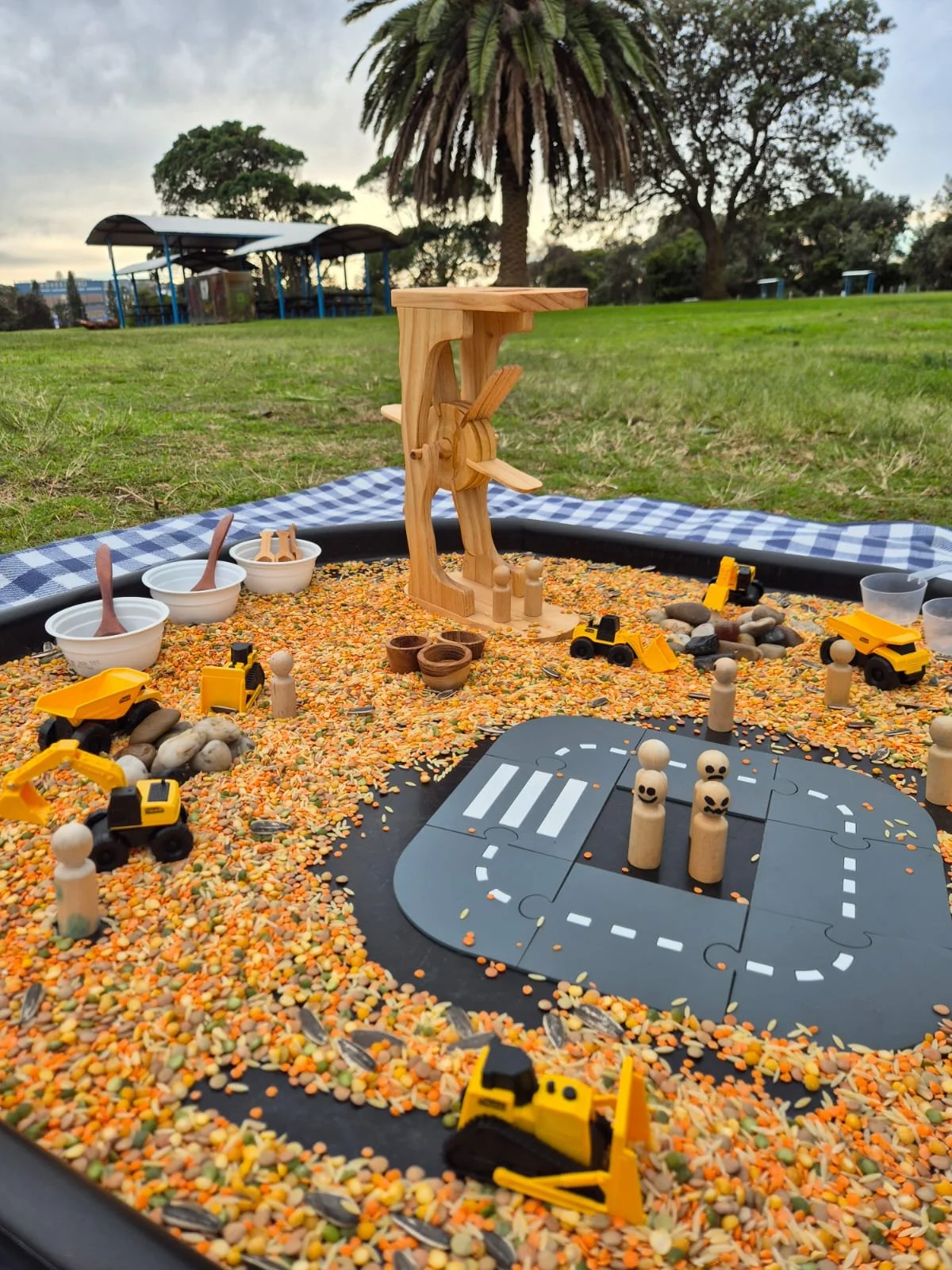 A sandbox with toy construction vehicles, wooden figures, and a wooden airplane in a park setting with trees and grass in the background.