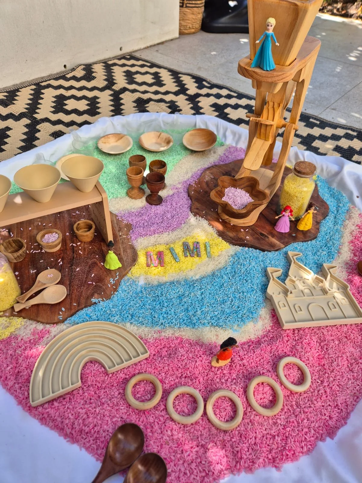 A colorful sand and rice sensory tray with wooden and plastic toys, including a castle mold, pink and purple princess figurines, a small castle print, a wooden slide, bowls, spoons, and small wooden cups, arranged on a rainbow-colored textured cloth.