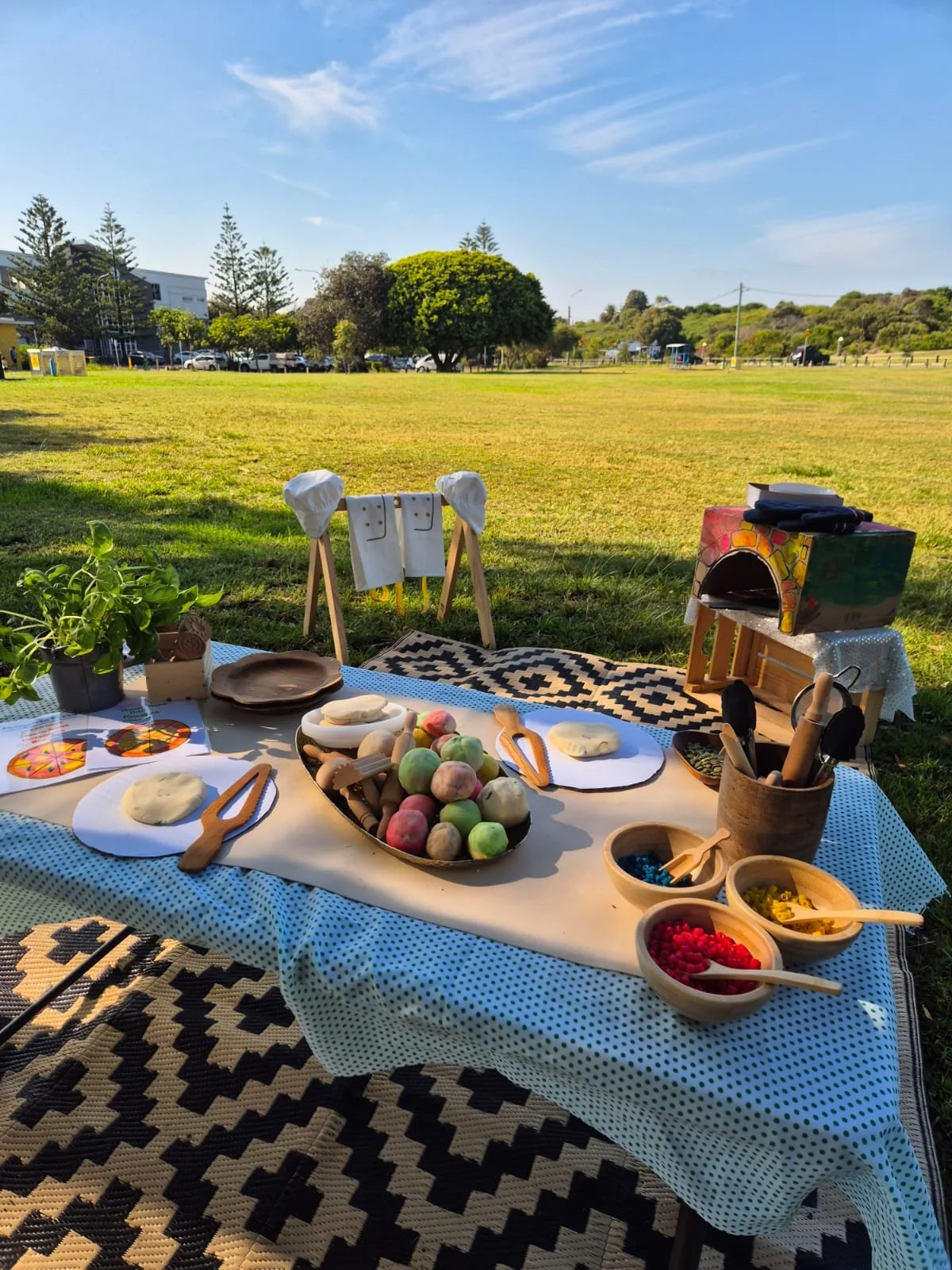 A colorful outdoor picnic setup with art supplies, clay, and toys on a table covered with a white and black patterned cloth, with a grassy field and trees in the background