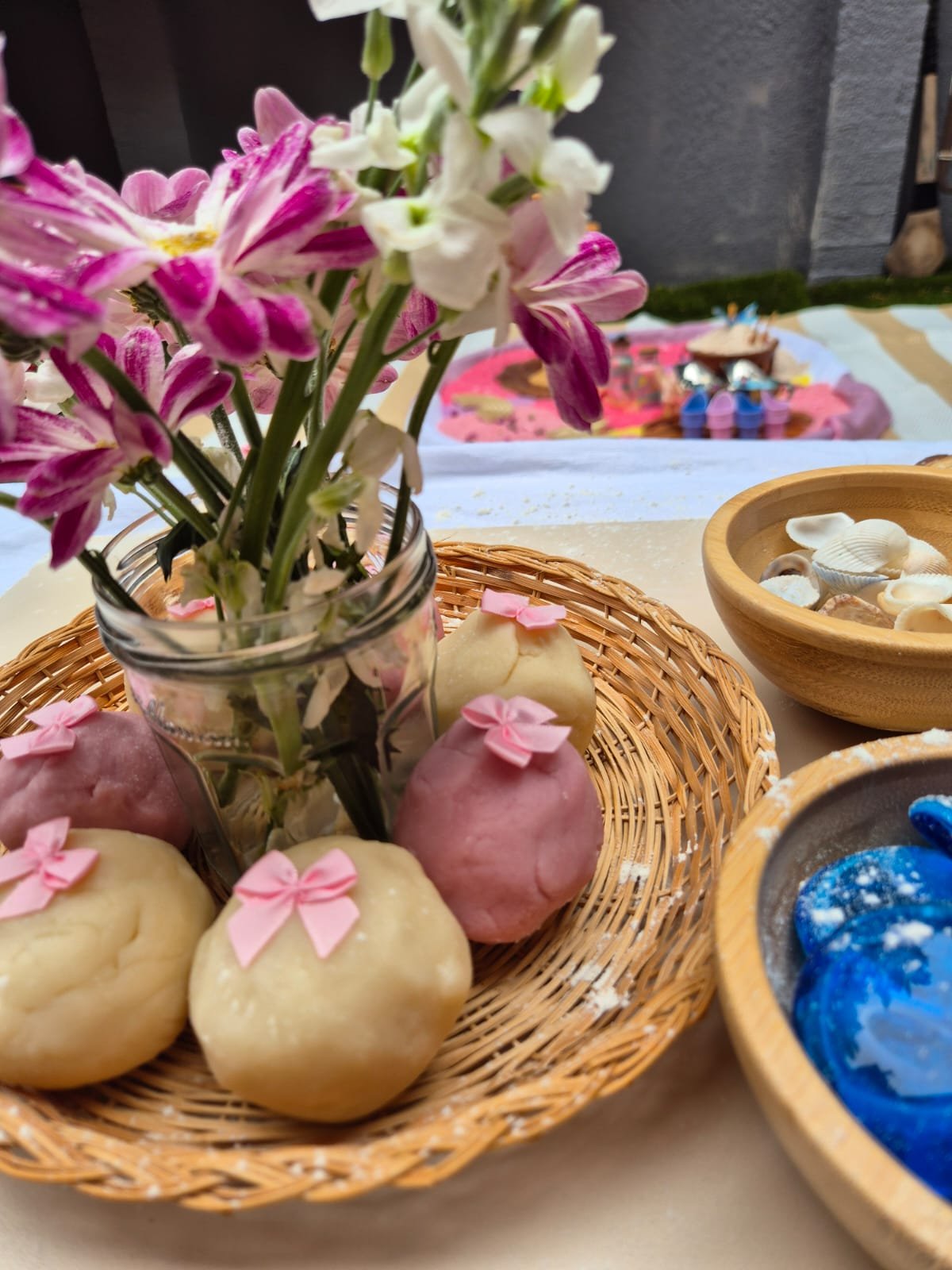A wicker basket with decorated cookies, a jar with pink and white flowers, and several bowls of seashell-shaped desserts on a table.