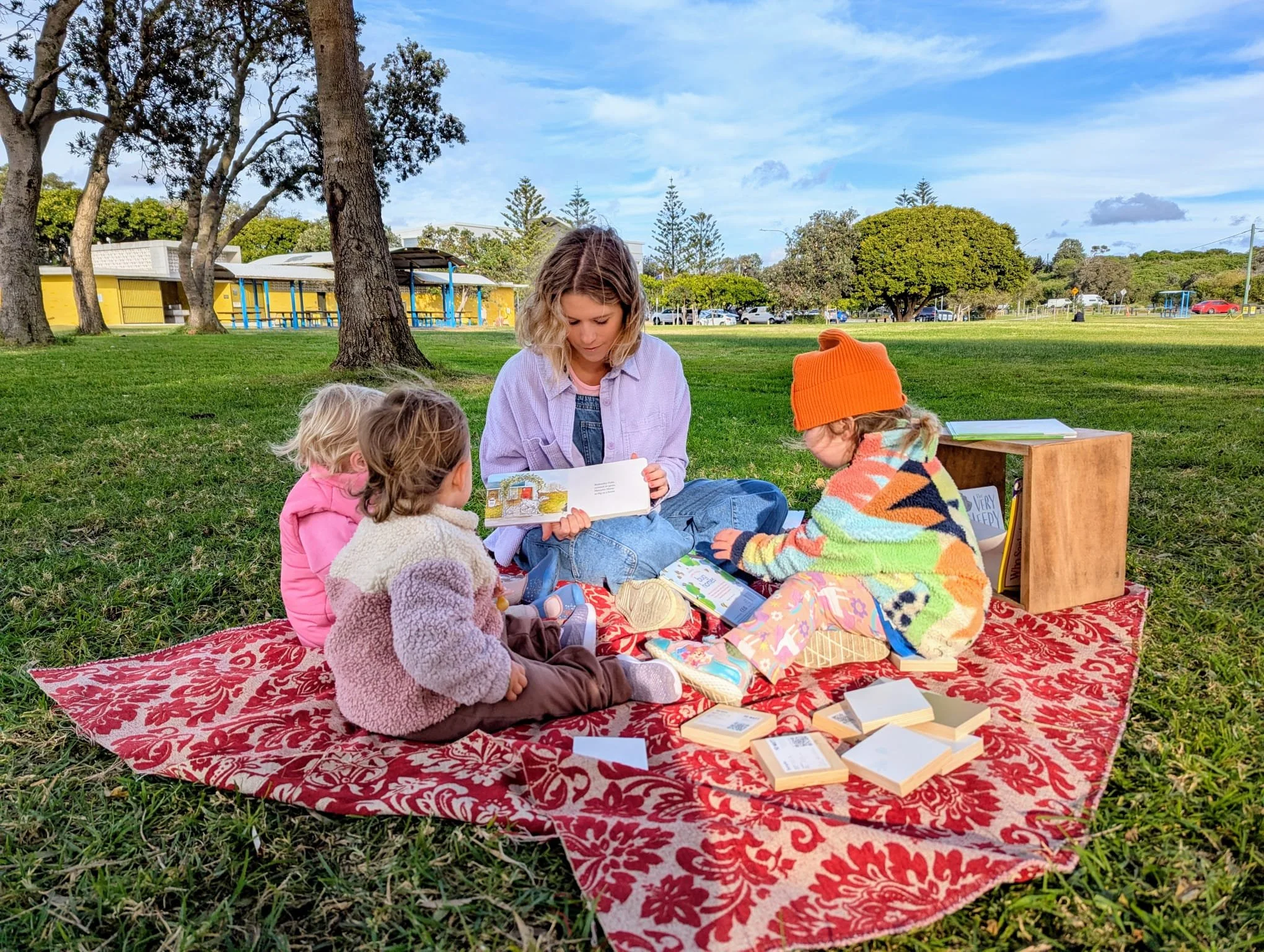 A woman sitting on a red patterned blanket reading a book to children in a park with trees, grass, and a yellow building in the background.