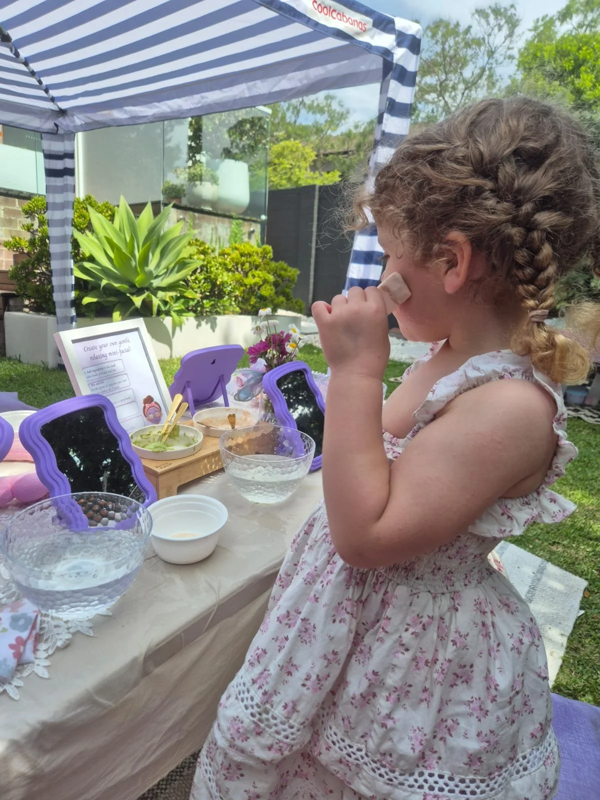 A young girl with curly hair in a braid is standing outdoors at a table with a purple mirror, bowls, and a sign about creating a mint facial, under a blue and white striped canopy. She is holding a tissue to her eye.